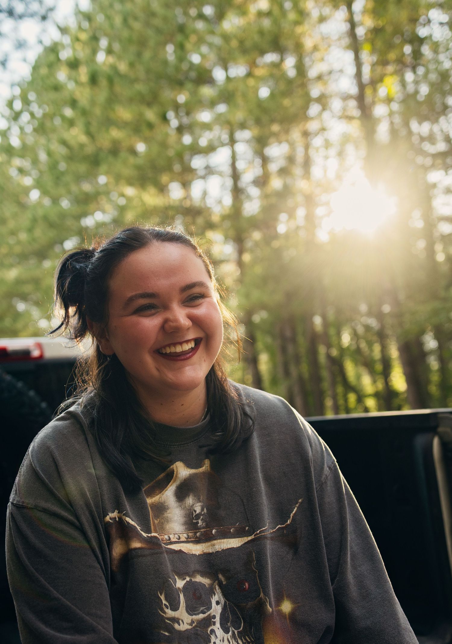 Smiling young woman in graphic sweatshirt laughing outdoors with sunlight streaming through forest trees.