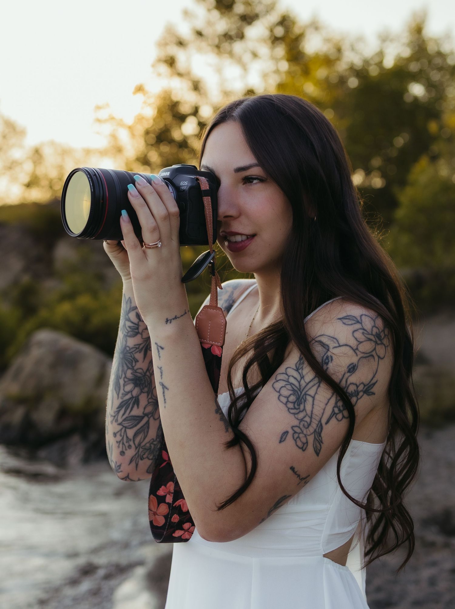 Tattooed woman with long dark hair holds a DSLR camera outdoors, wearing a white dress at golden hour.