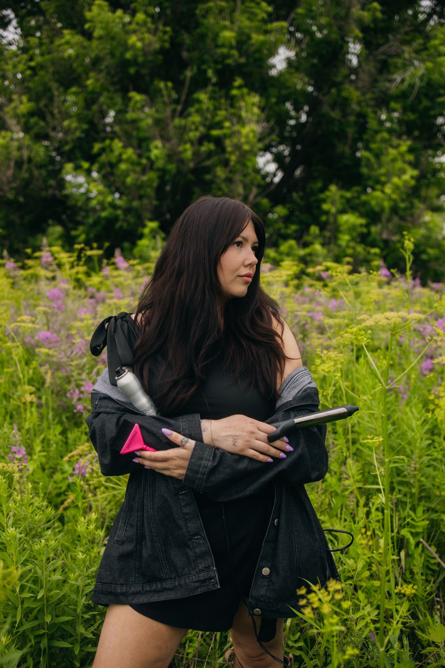 Woman in black jacket holding hair tools in a lush green wildflower field.