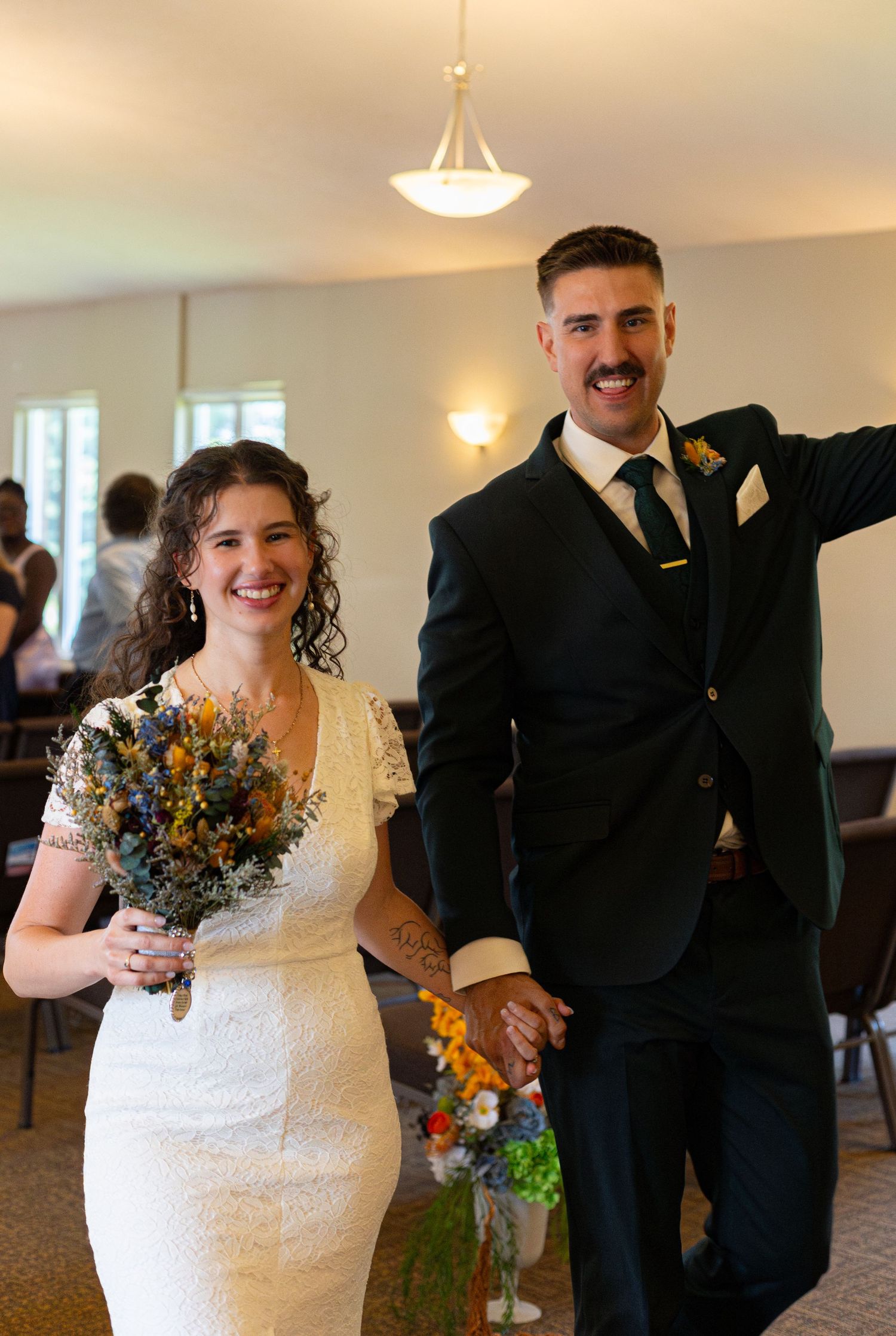 Smiling bride in white lace dress holding wildflower bouquet stands with groom in dark suit at wedding ceremony.