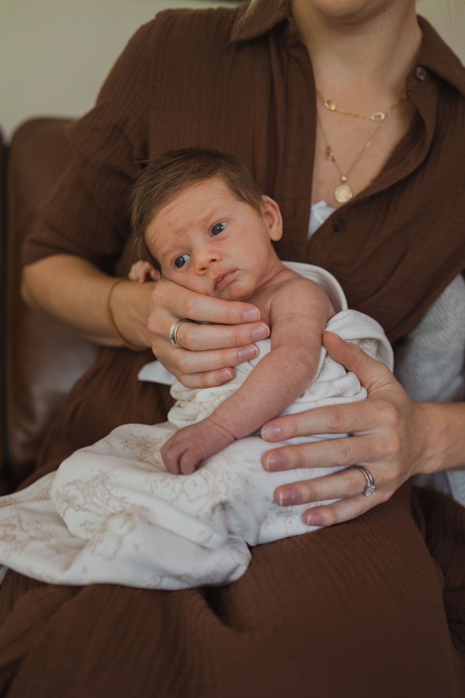 A mother in a brown dress gently holds her newborn baby wrapped in a white blanket.