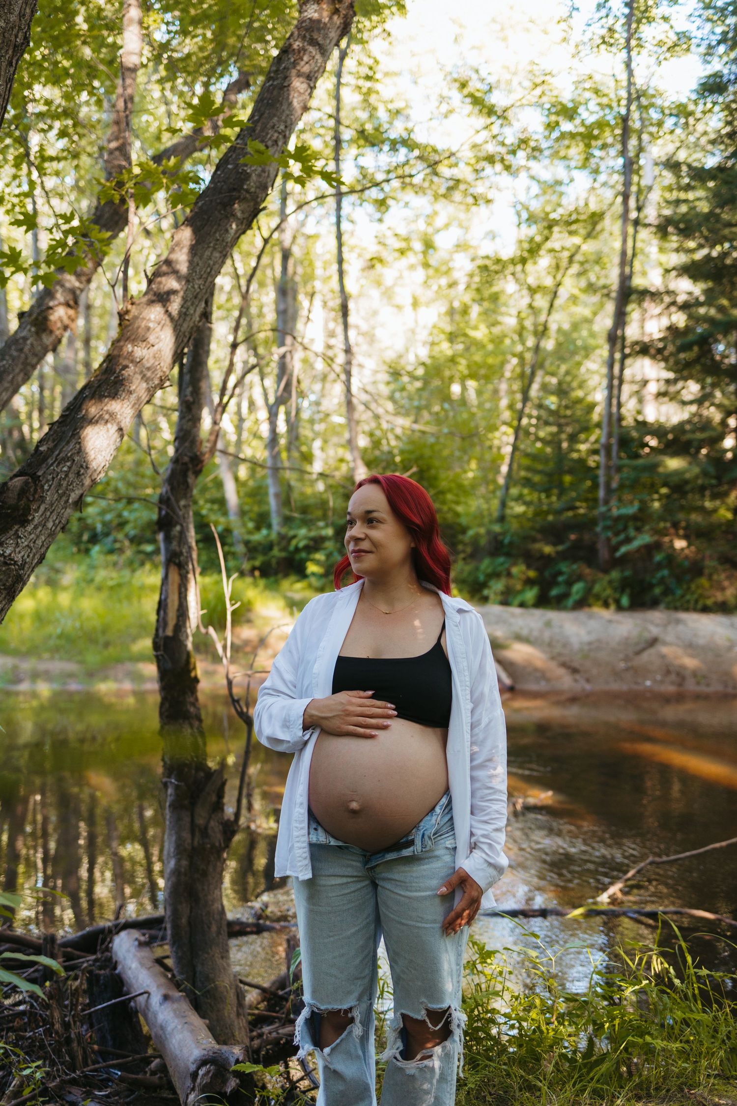 Pregnant woman with red hair cradles her belly outdoors by a forest creek, wearing ripped jeans and white shirt.