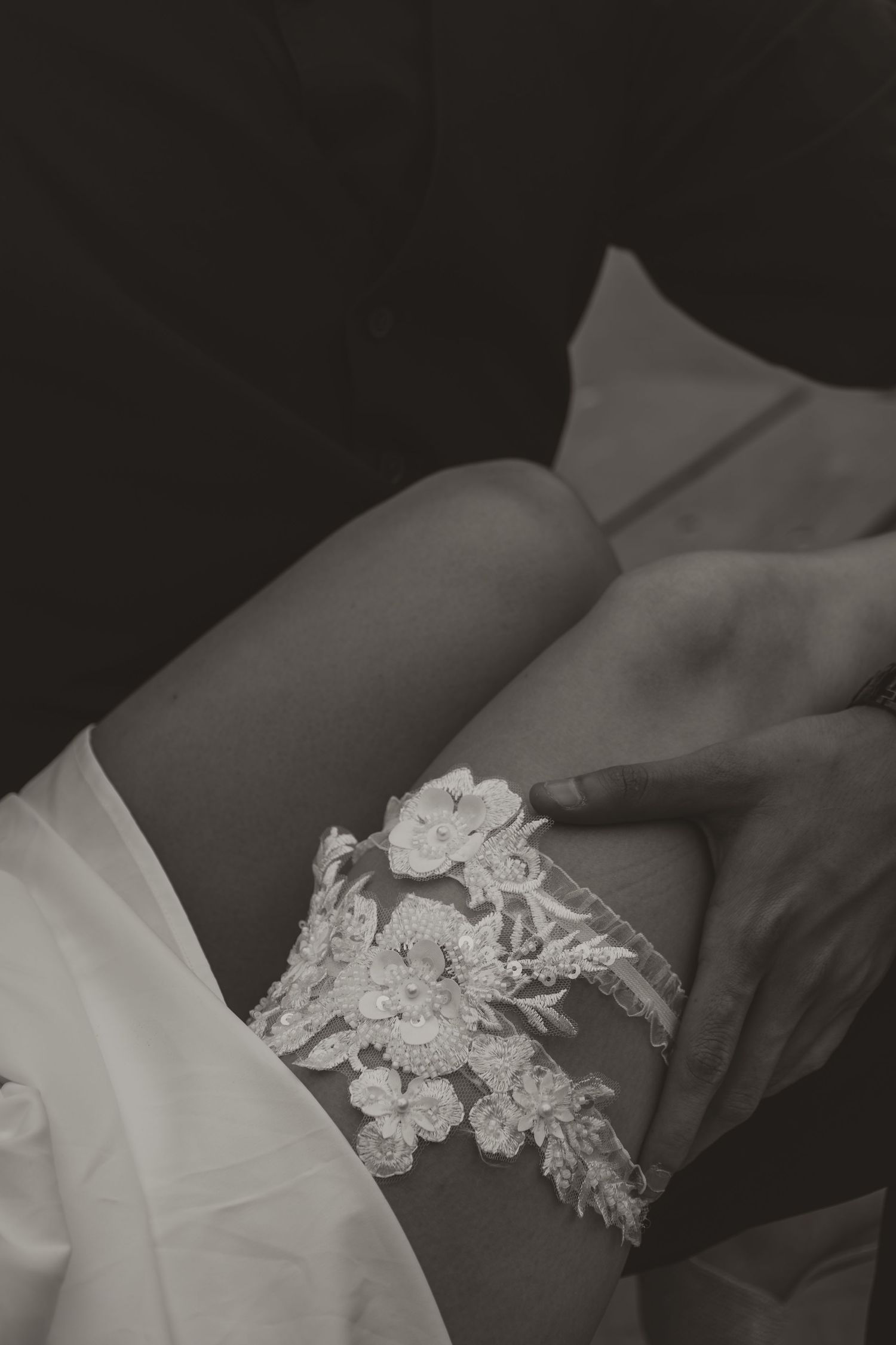 Black and white close-up of bride placing lace floral garter on leg during wedding preparations.