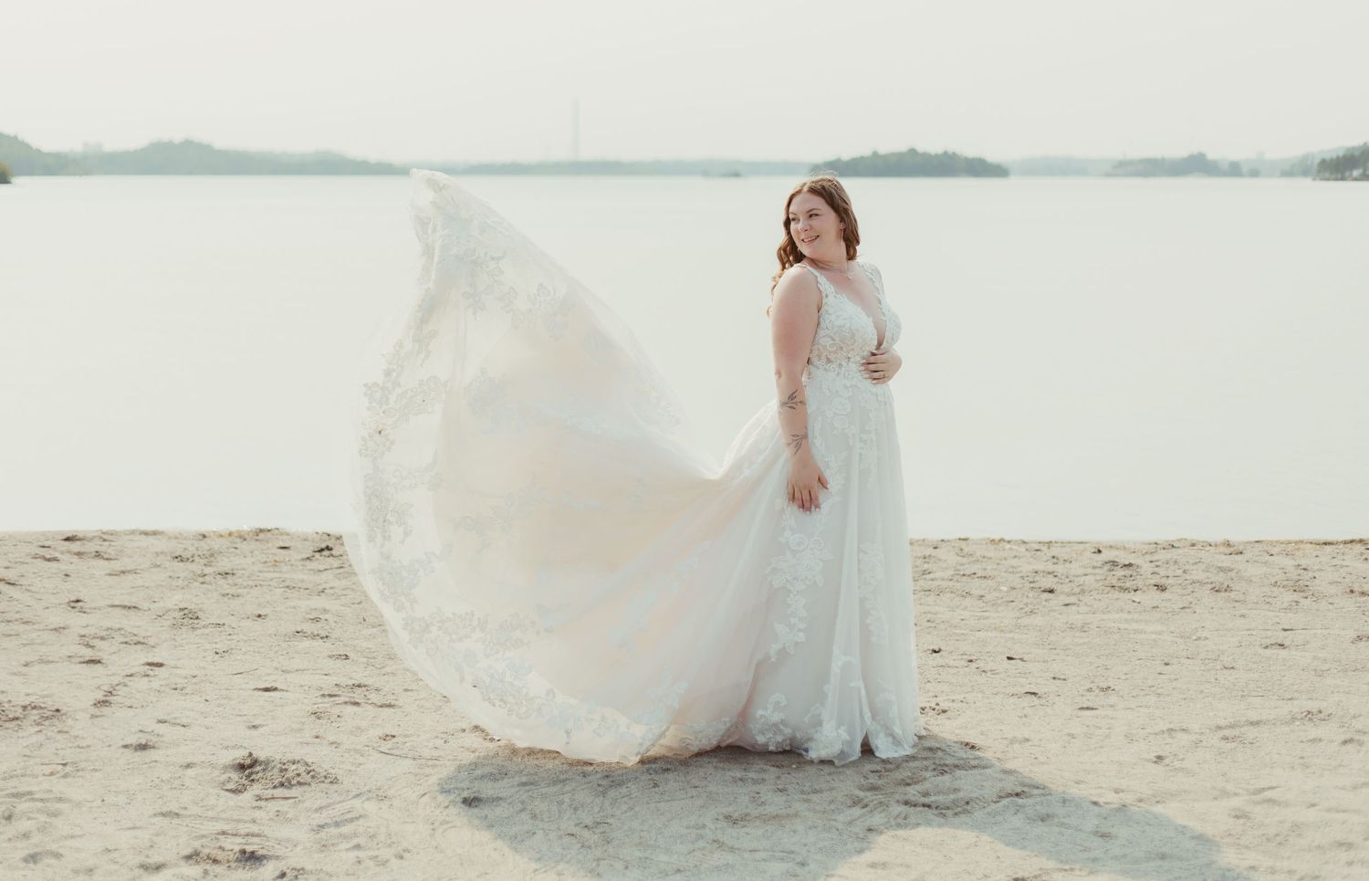 Bride in flowing white lace gown with veil billowing in wind on sandy lakeside beach.