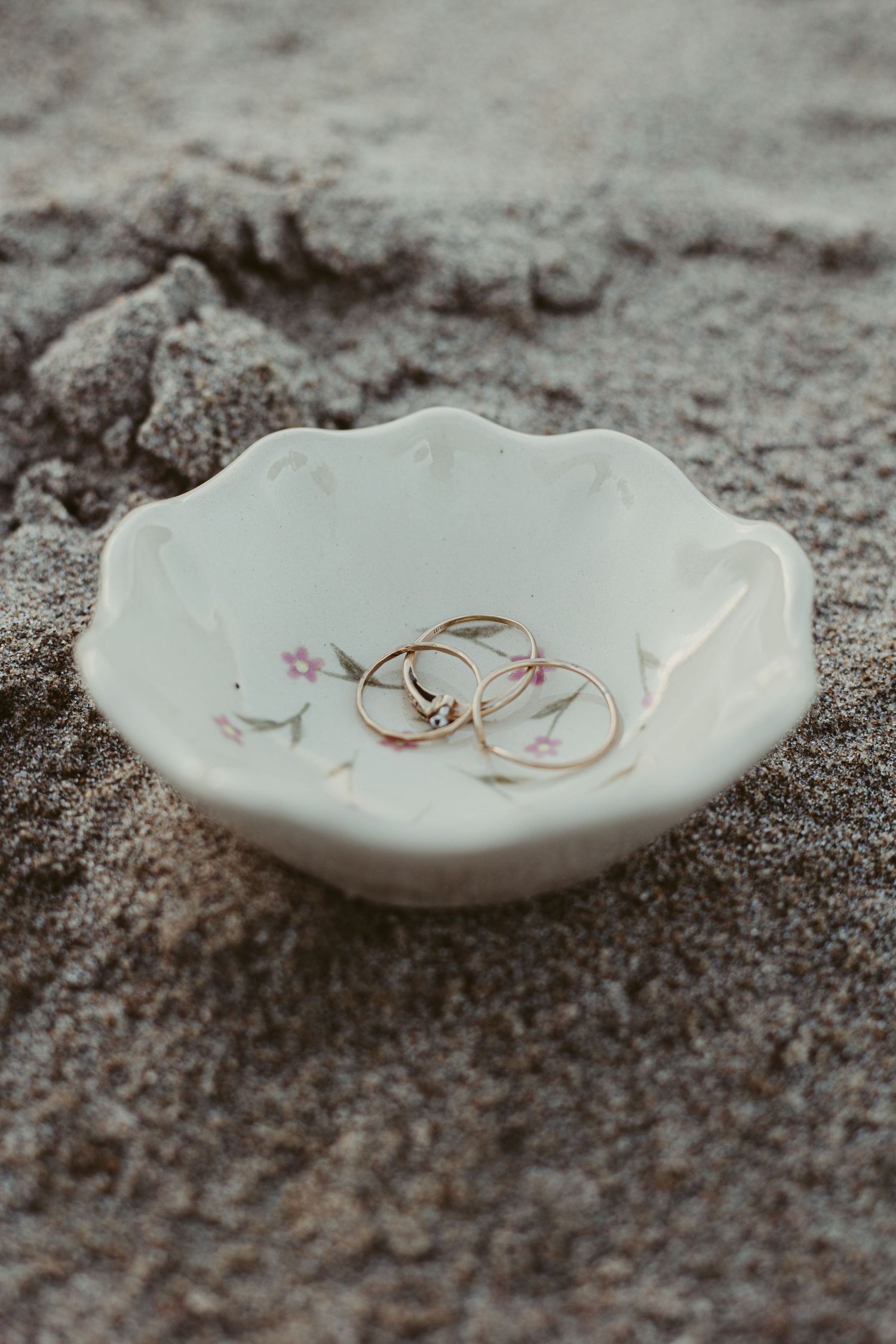 Gold wedding rings and diamond ring displayed in white floral ceramic dish on sandy beach.