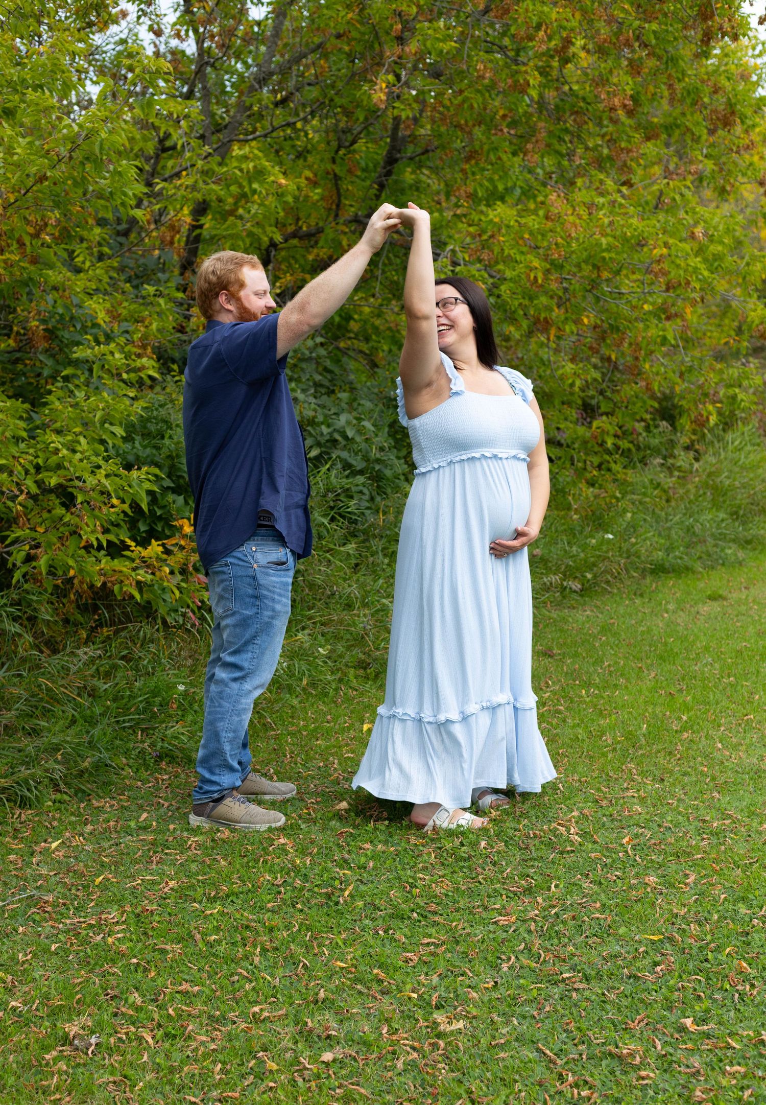 Expecting couple dancing outdoors during maternity photoshoot, woman in flowing light blue dress holding baby bump.