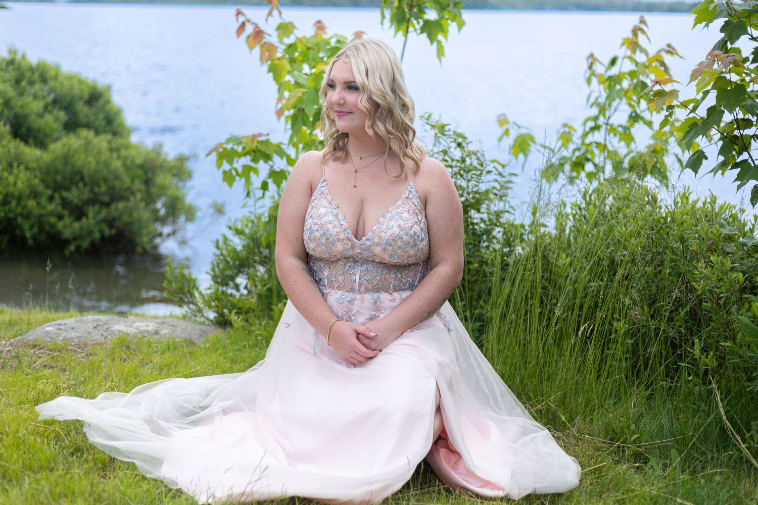 A blonde woman in a pink beaded gown sits gracefully on grass beside a serene lakeside setting.