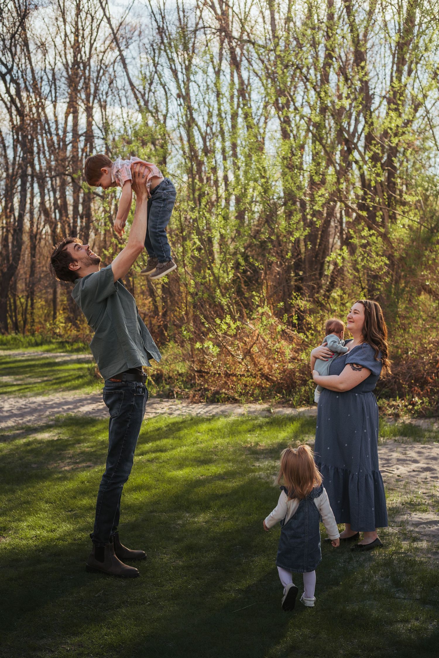 Family of four enjoying a sunny outdoor moment, dad tossing toddler in the air while mom holds baby near lush green trees.