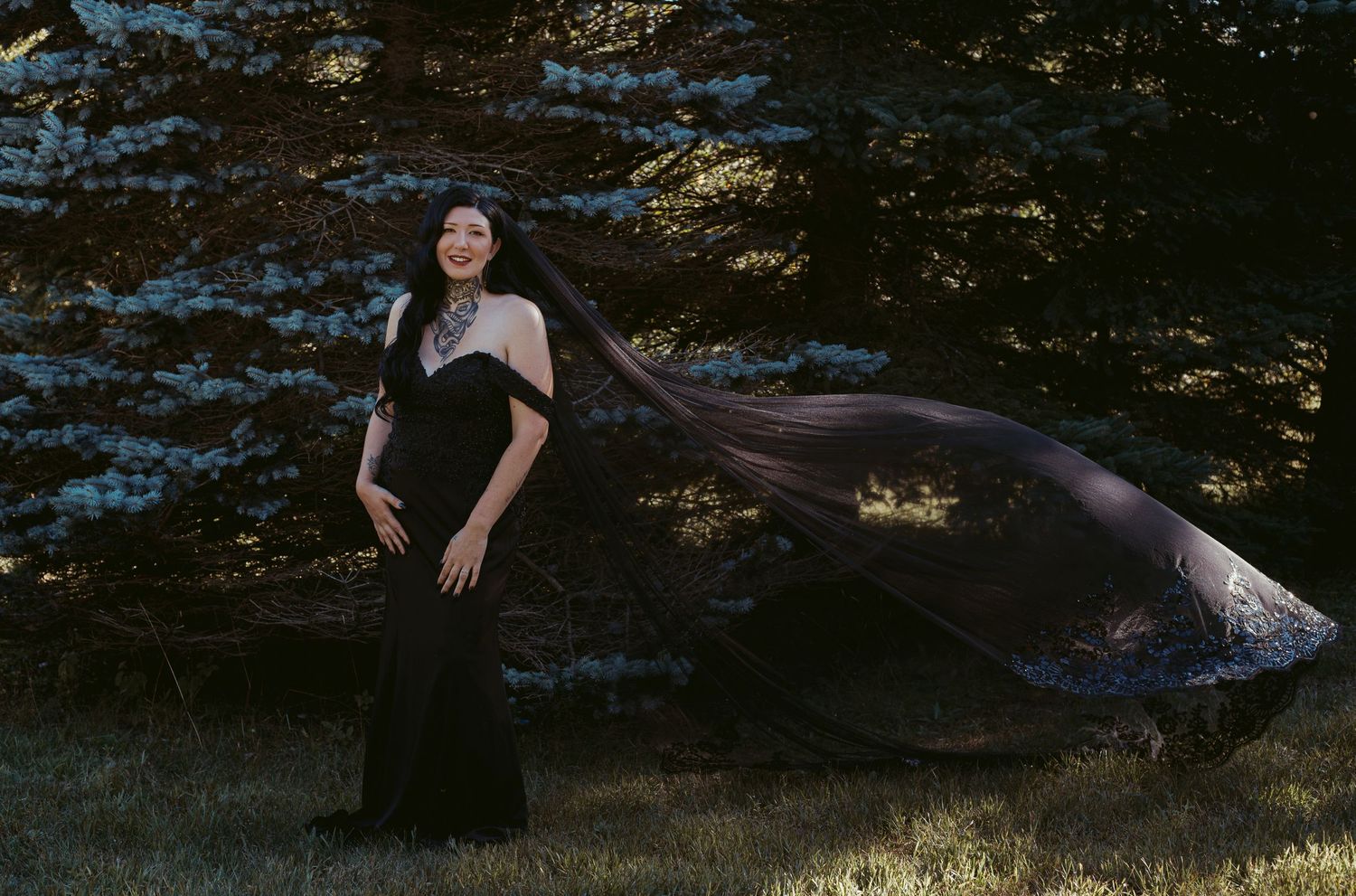 Woman in elegant black gown with flowing cape standing gracefully among trees in golden light.