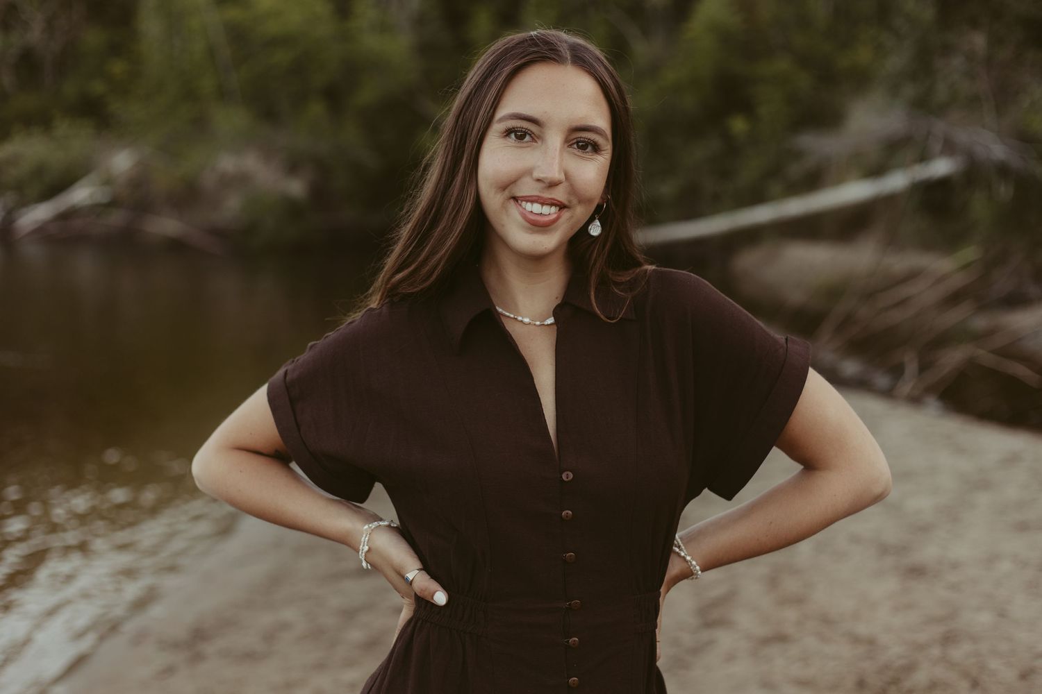 Smiling woman in black button-up dress posing confidently outdoors near a river.
