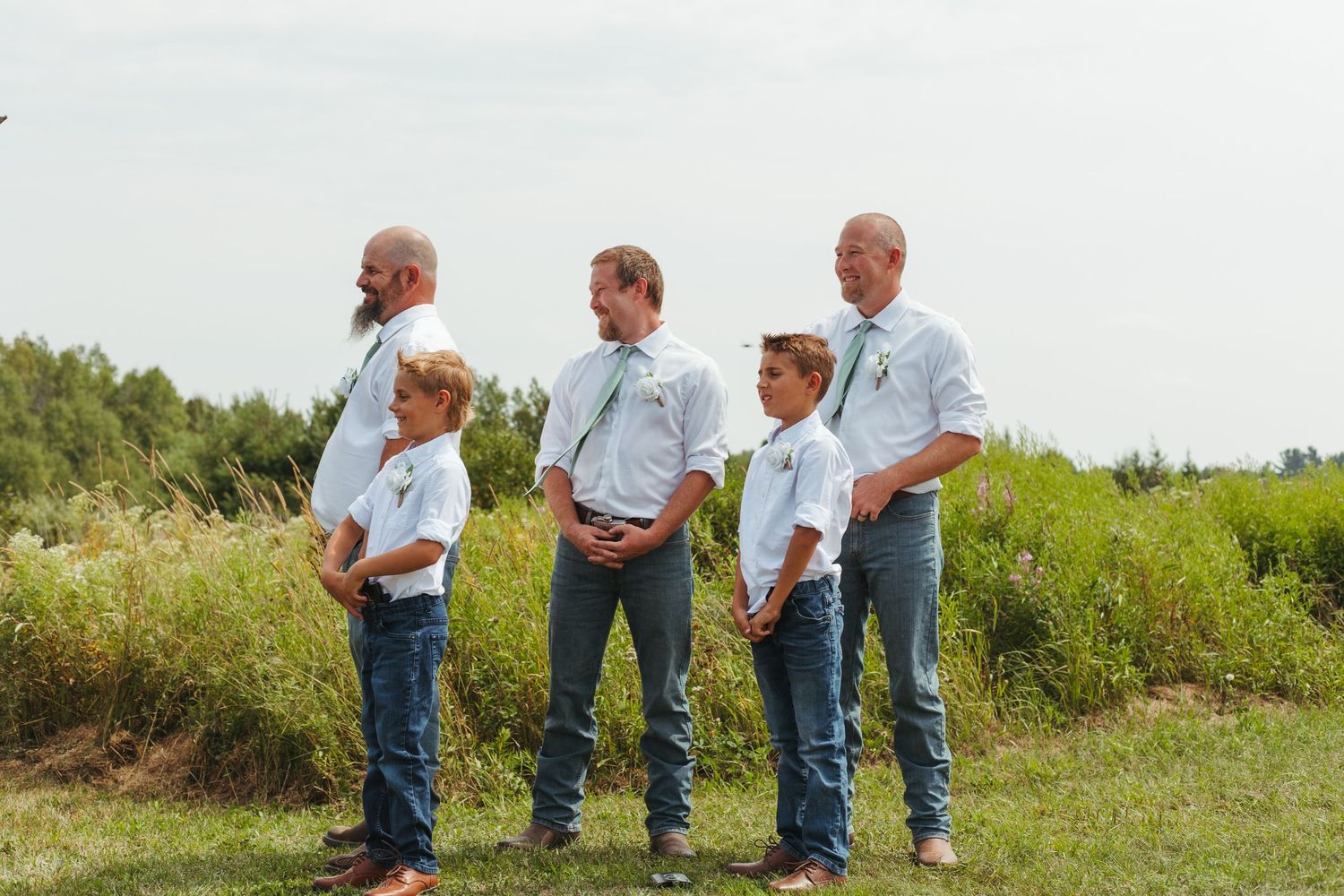 Groomsmen and ring bearers in white shirts and jeans posing outdoors at wedding.