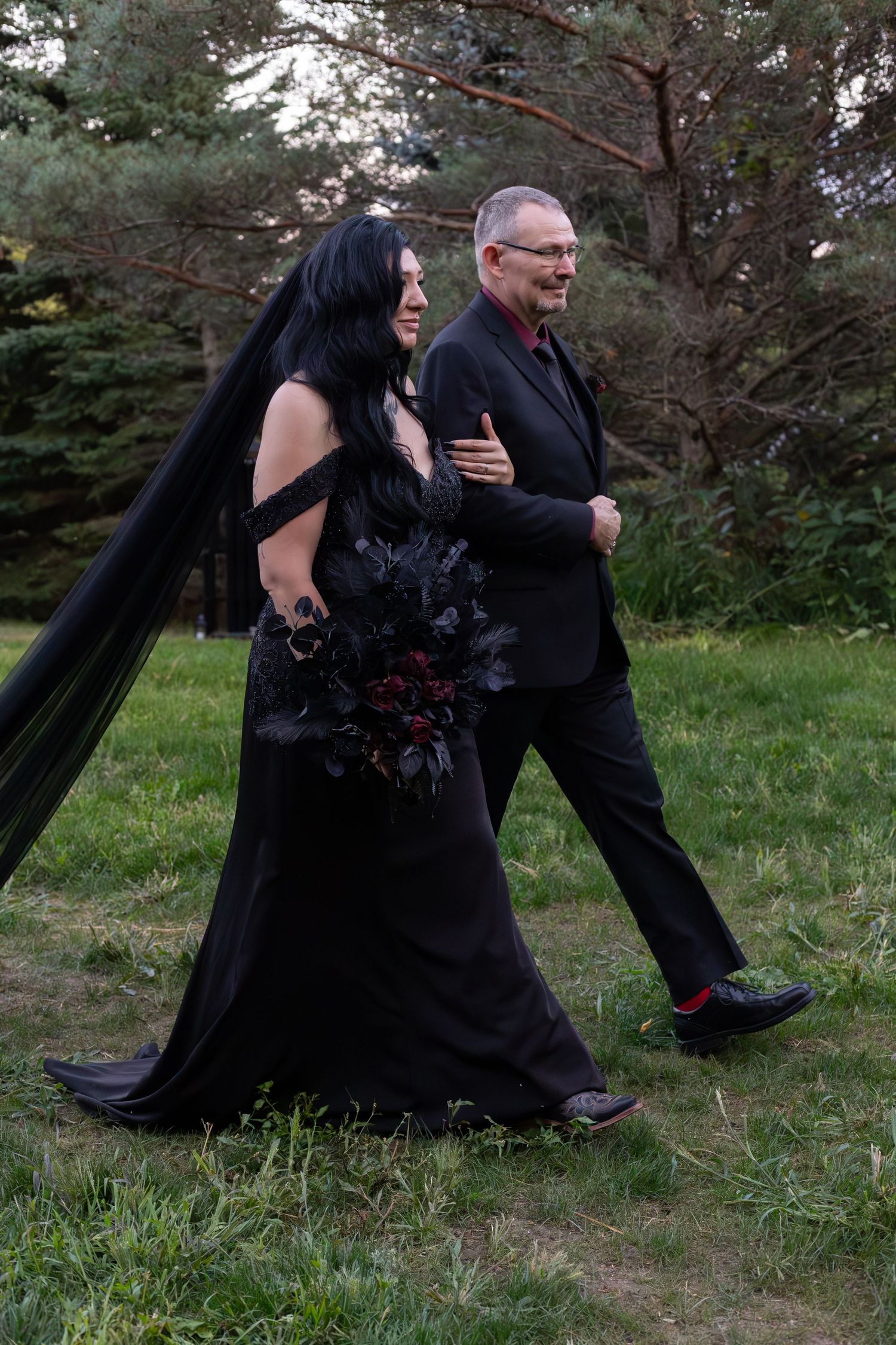 Bride in black gown with dark veil and bouquet walks with groom through a forest wedding ceremony.