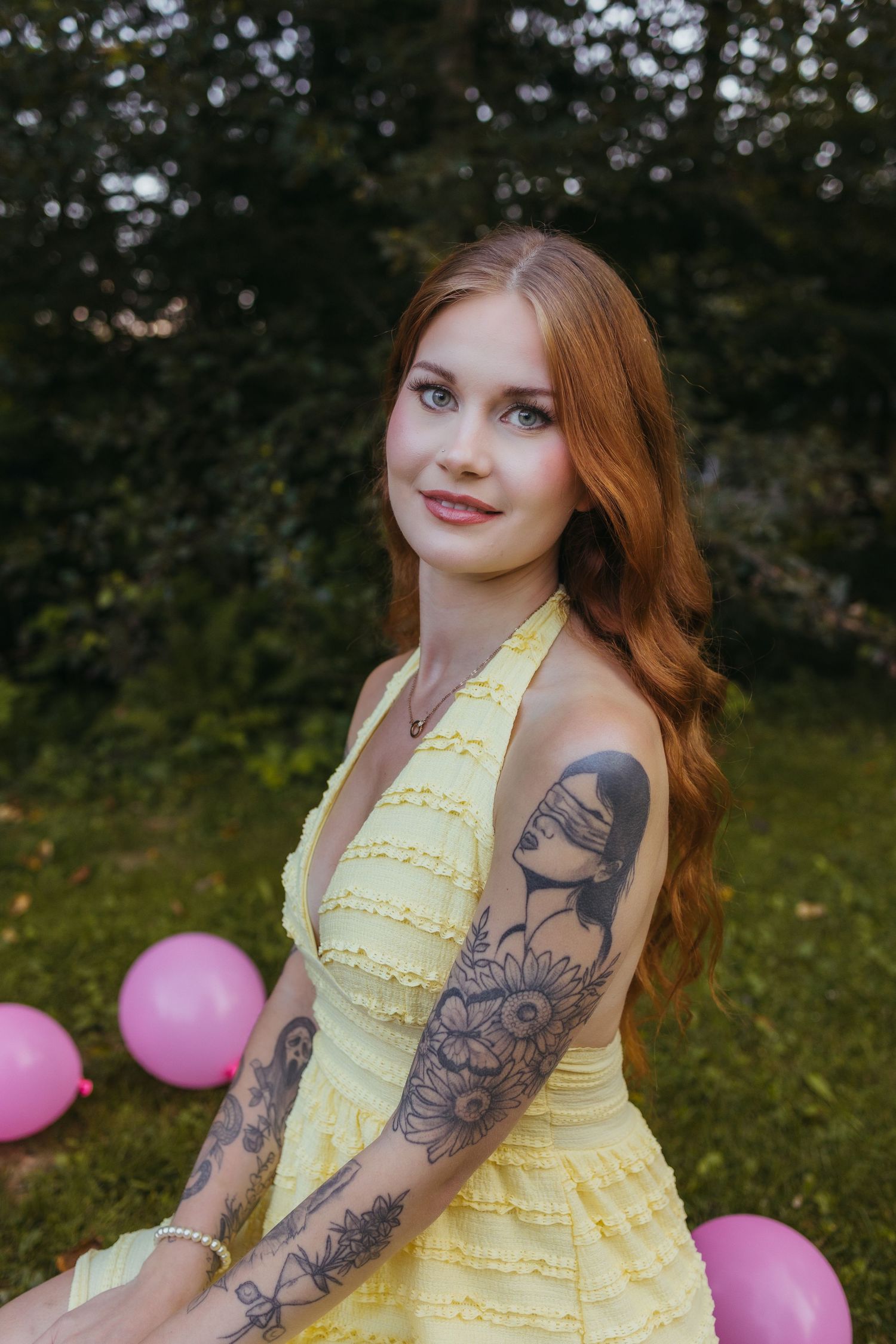 Young woman with red hair and tattoos in yellow halter dress sits outdoors with pink balloons.