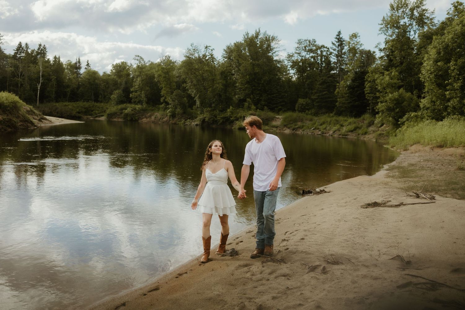Couple holding hands walking along sandy riverbank with lush green trees and calm water in background.