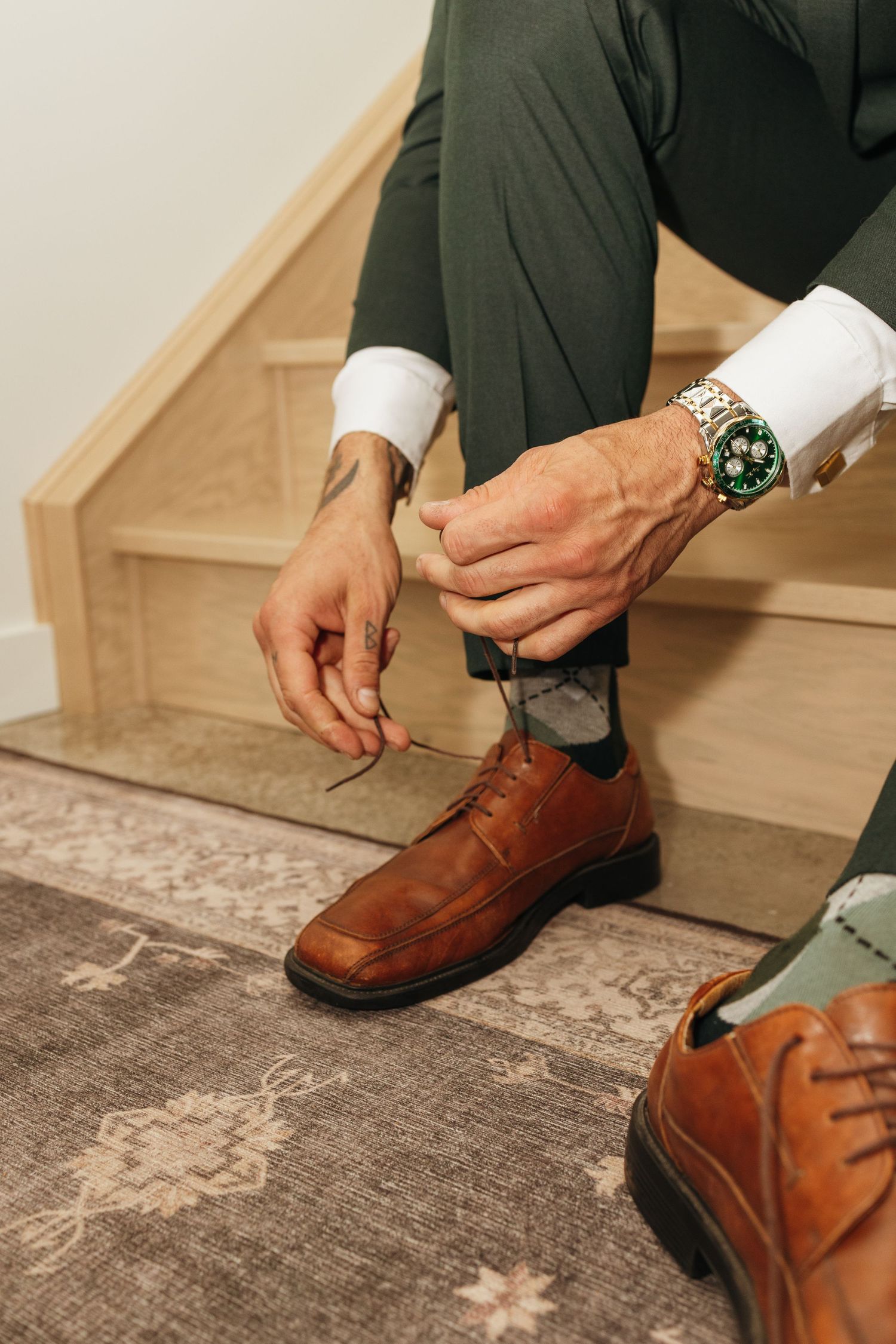 Man in green trousers and white shirt tying brown leather dress shoes on wooden stairs while wearing a luxury watch.