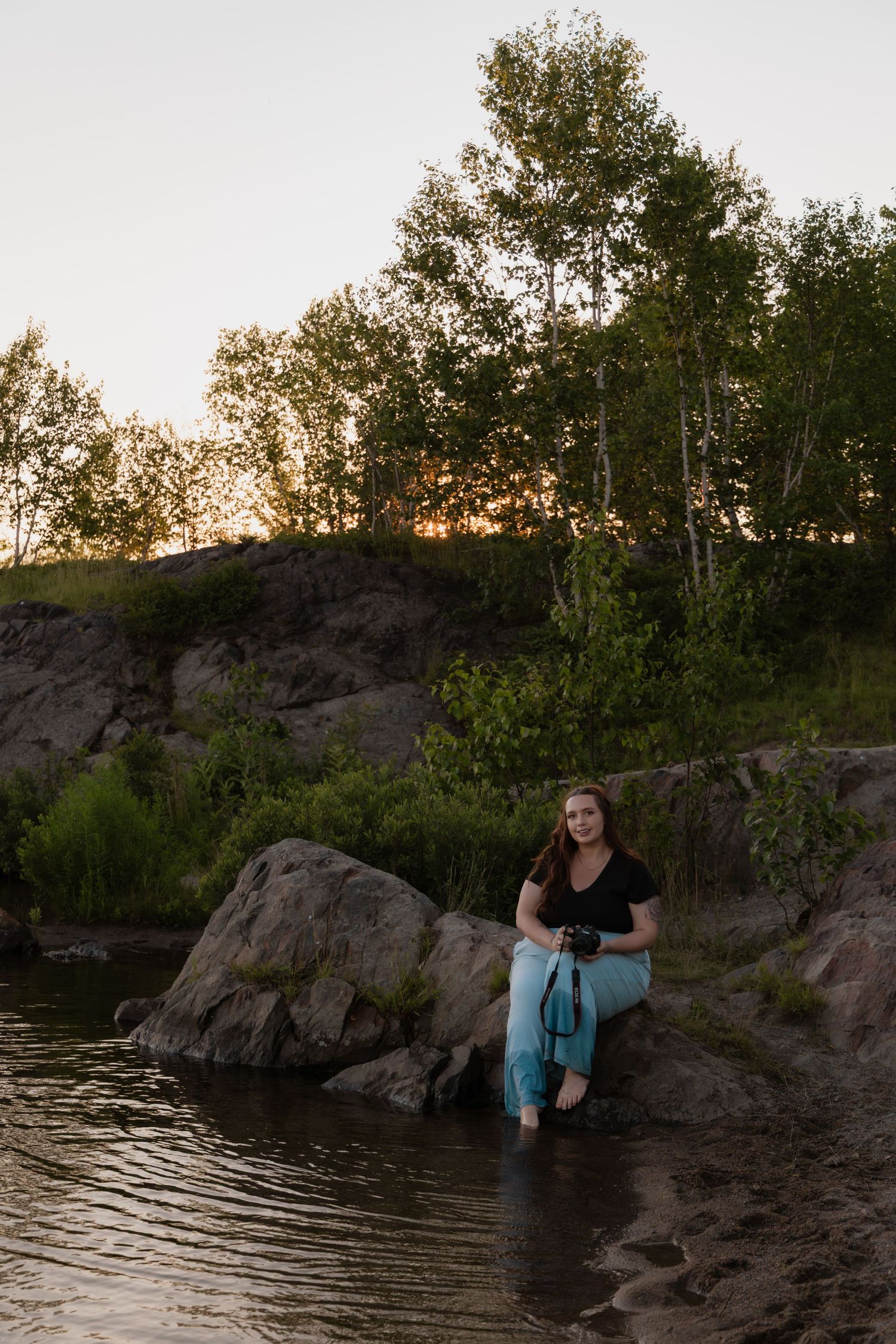 Woman sitting on rocks by a river at sunset, wearing black top and blue jeans, holding a camera, surrounded by trees.
