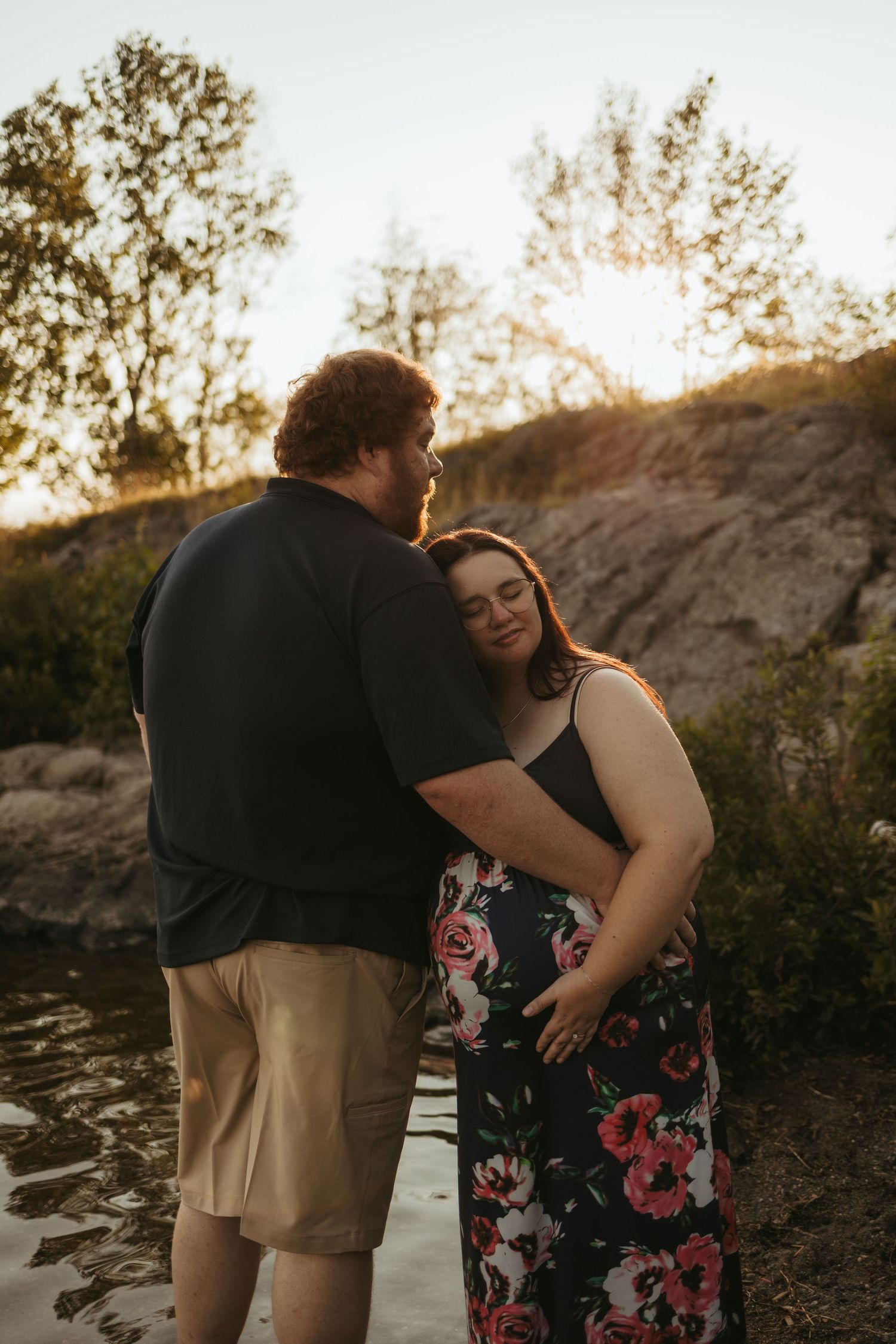 Couple sharing a tender moment outdoors at golden hour, woman in floral dress leaning on man near a rocky waterside.