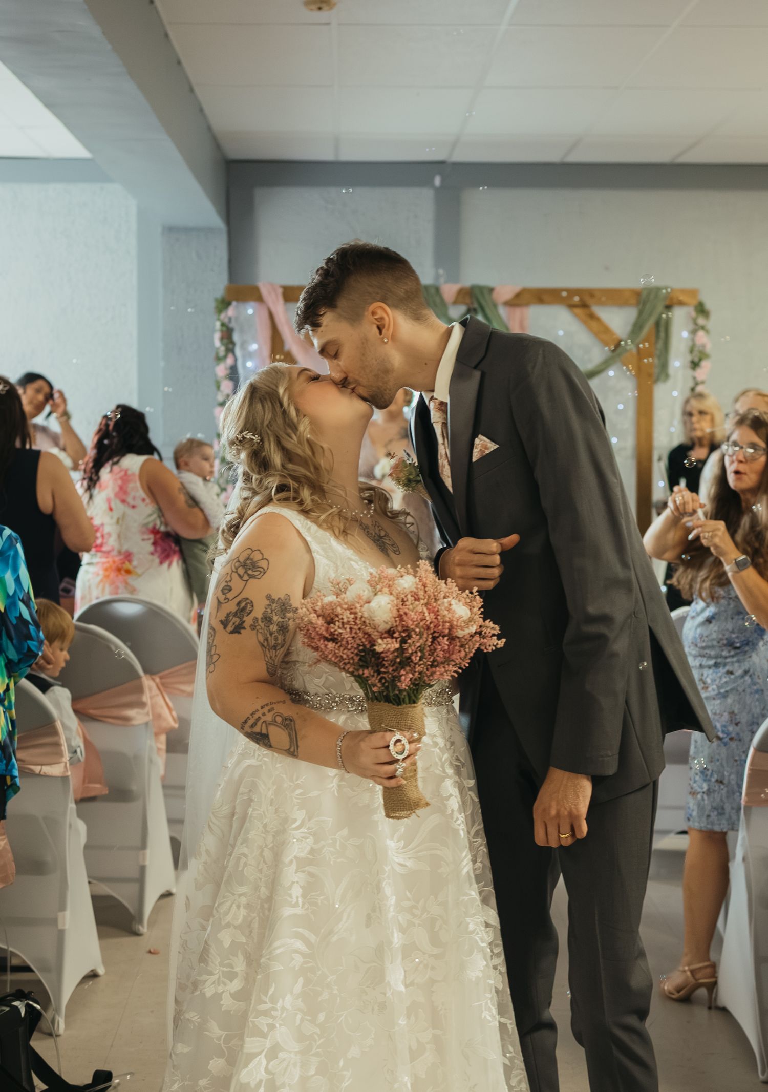 Newlywed couple shares first kiss in wedding ceremony aisle, bride in lace gown holding pink bouquet, groom in gray suit.