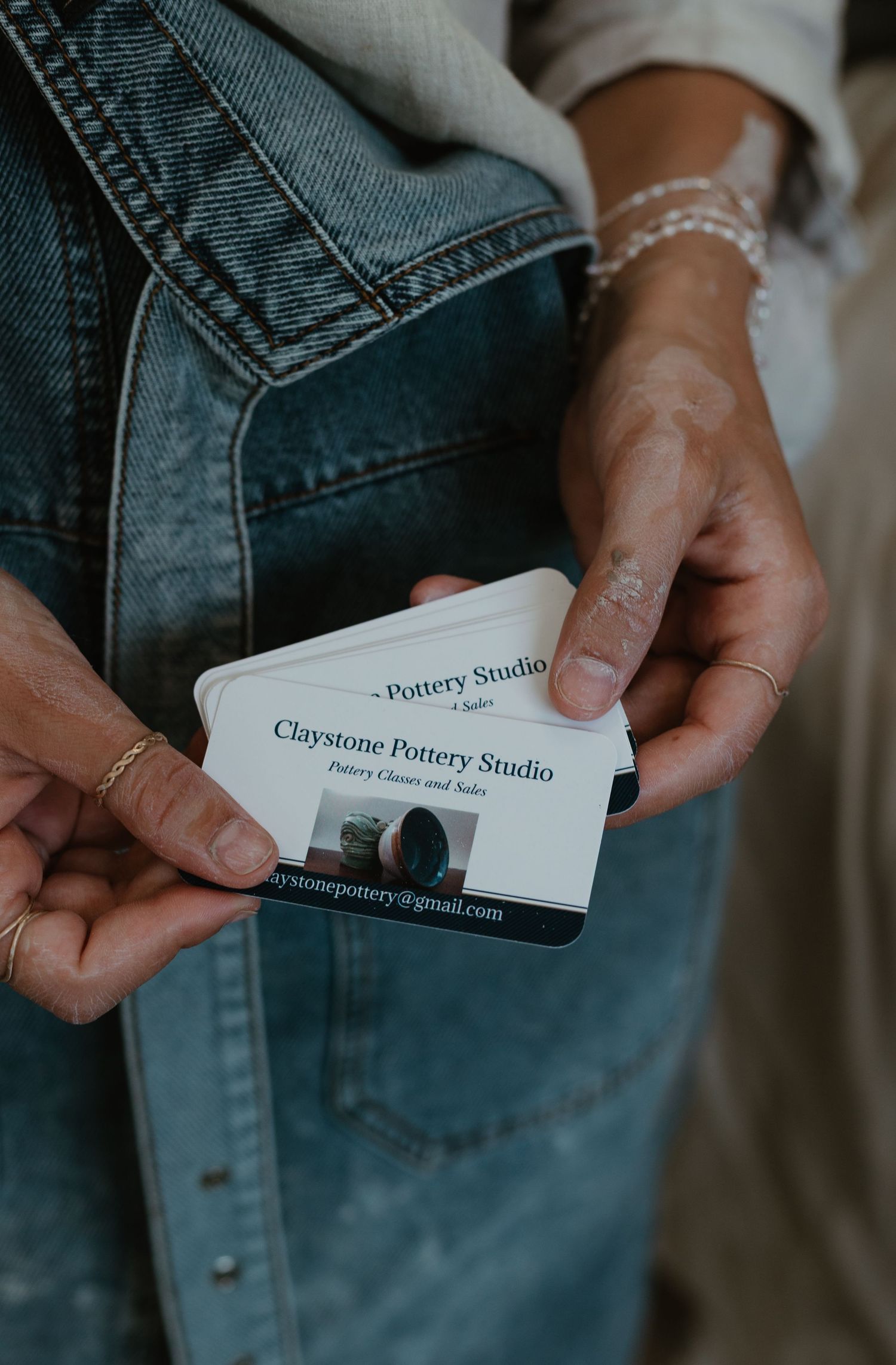 Hands holding Claystone Pottery Studio business cards, wearing gold rings and a bracelet, against a denim jacket.