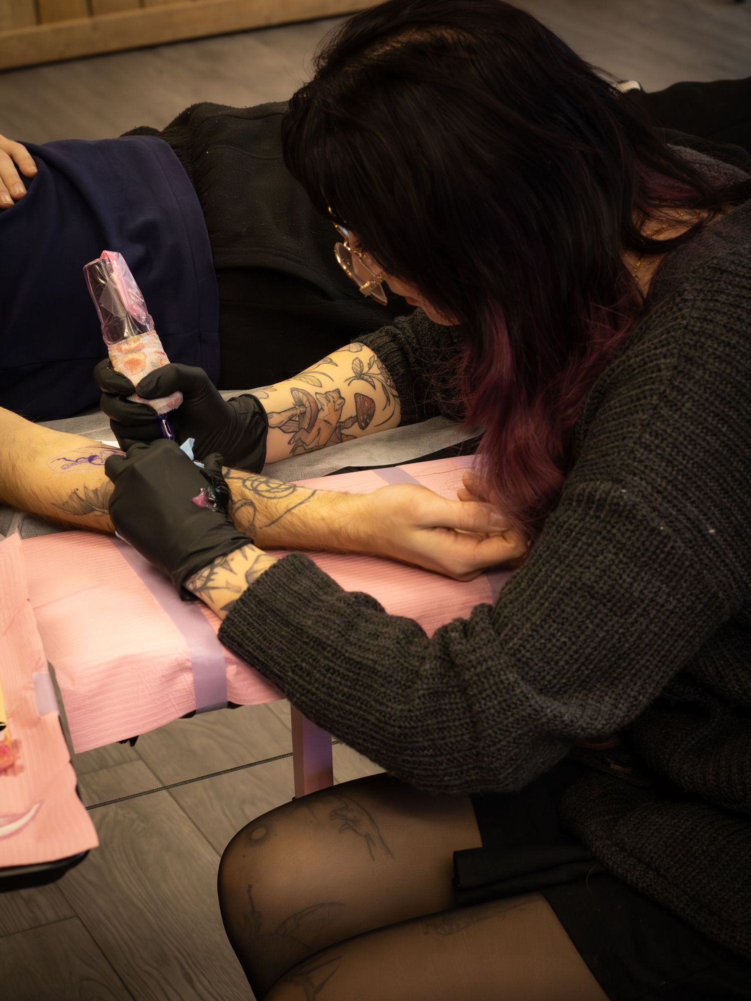 Tattoo artist with tattooed arms applying ink to a client's arm in a studio.