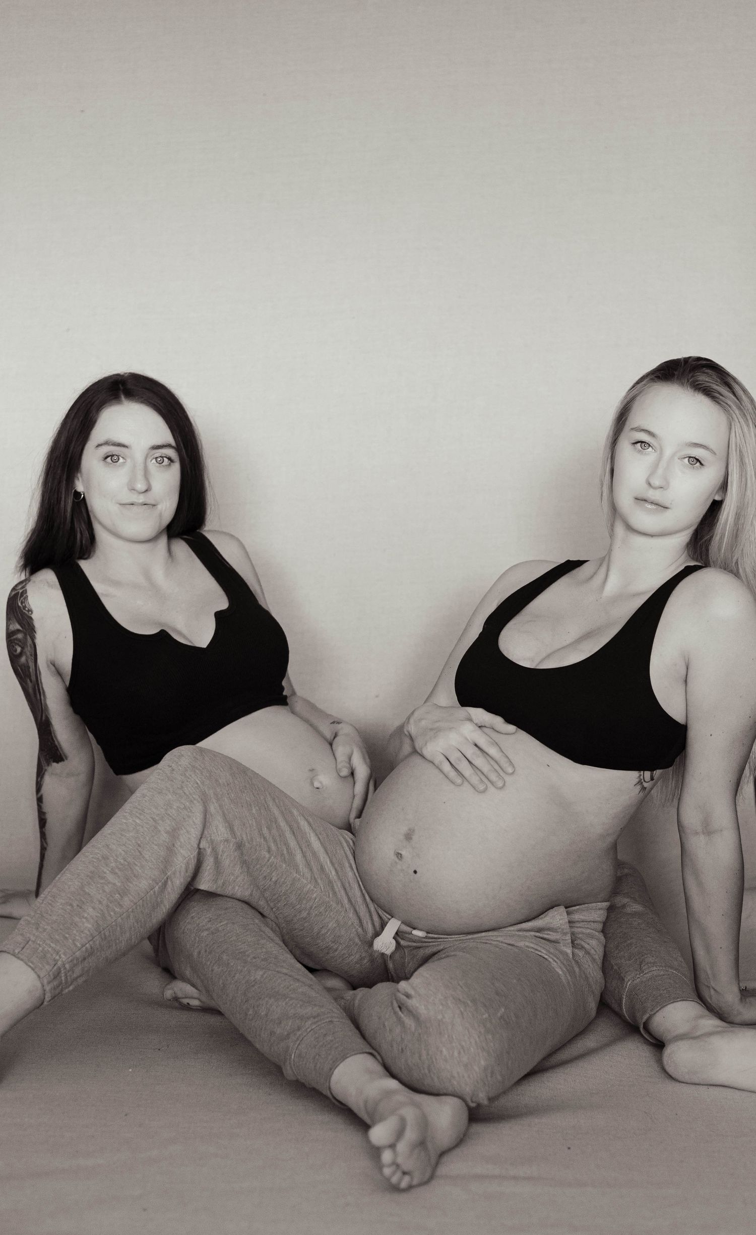 Black and white portrait of two pregnant women in black sports bras sitting together on bed.
