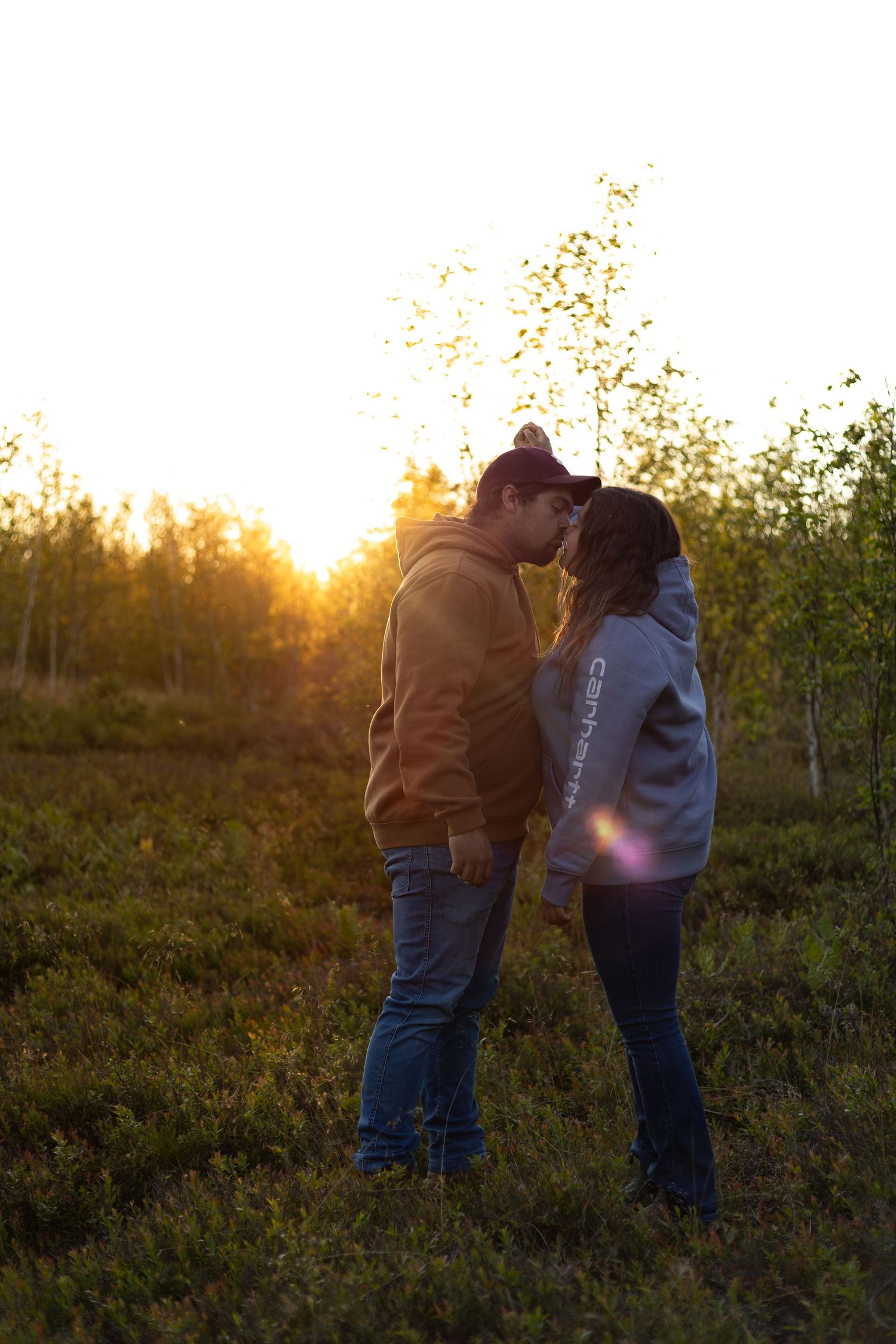 A couple shares a romantic kiss backlit by a glowing golden sunset in a field.