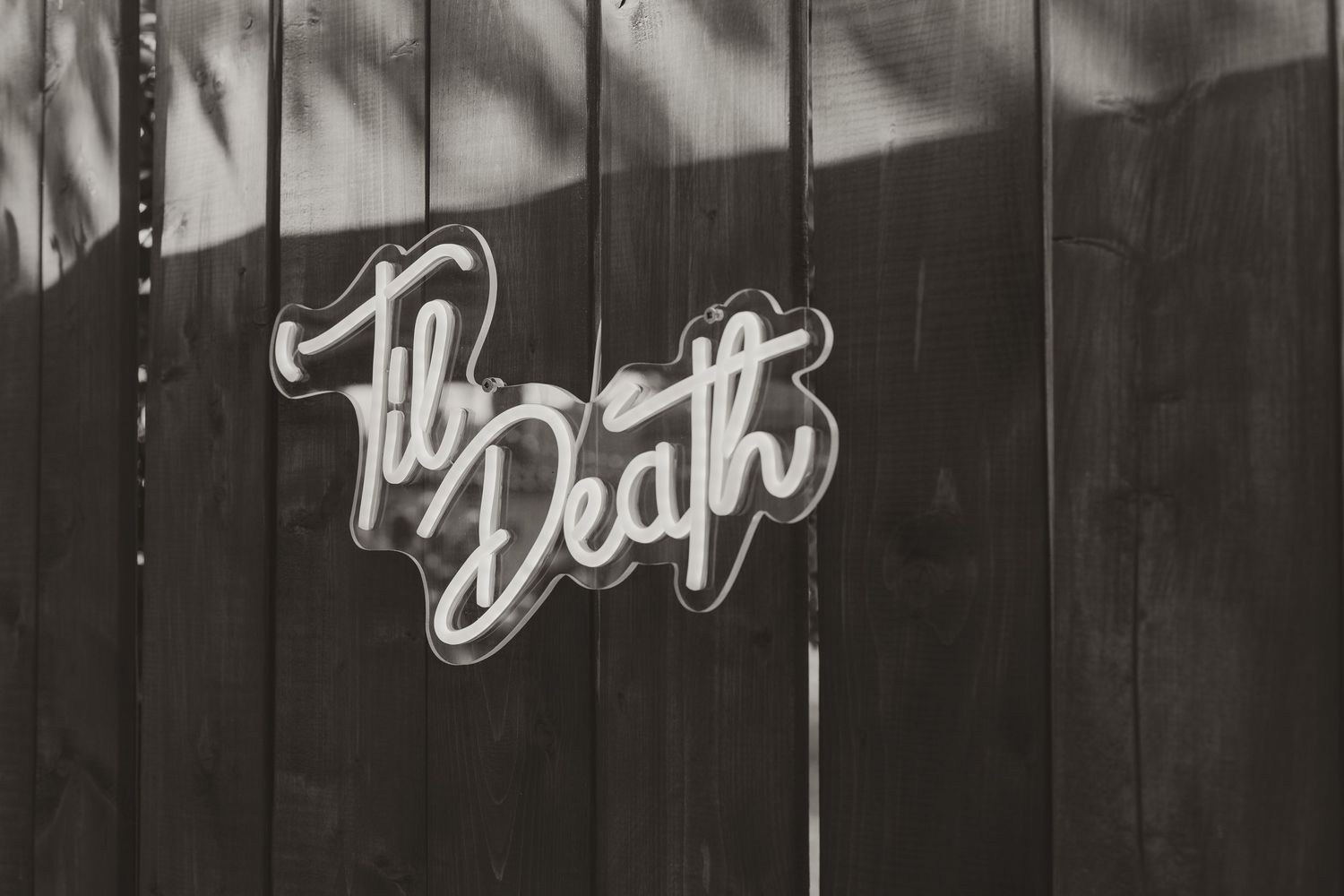 Black and white photo of decorative Til Death neon sign mounted on a dark wooden fence.
