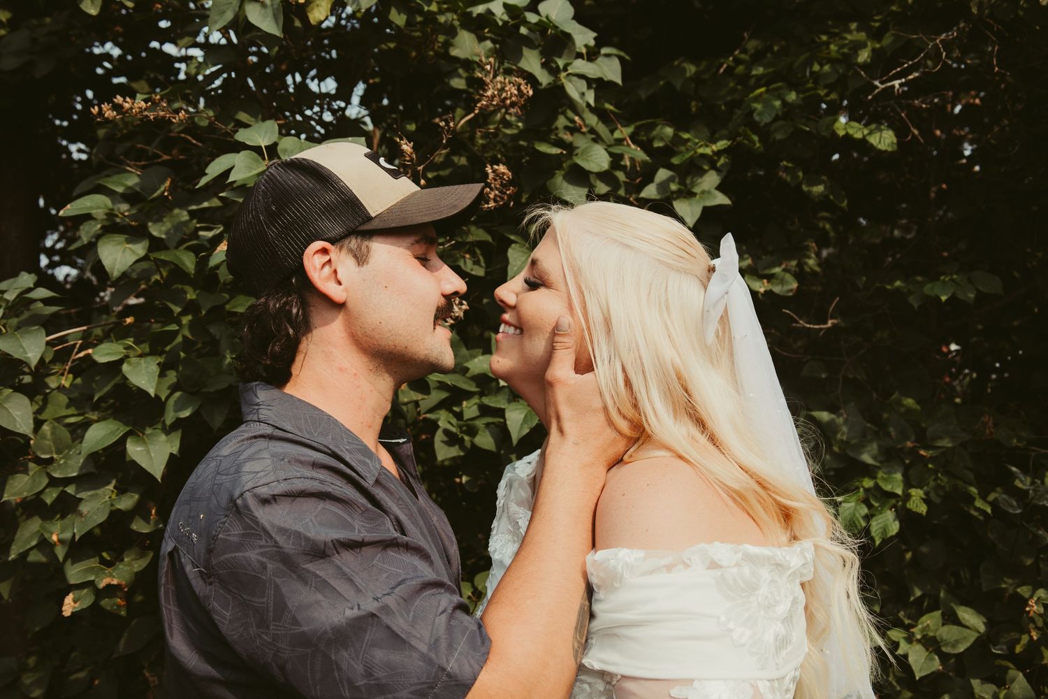 Groom in cap tenderly holding brides face as they share romantic moment surrounded by greenery.