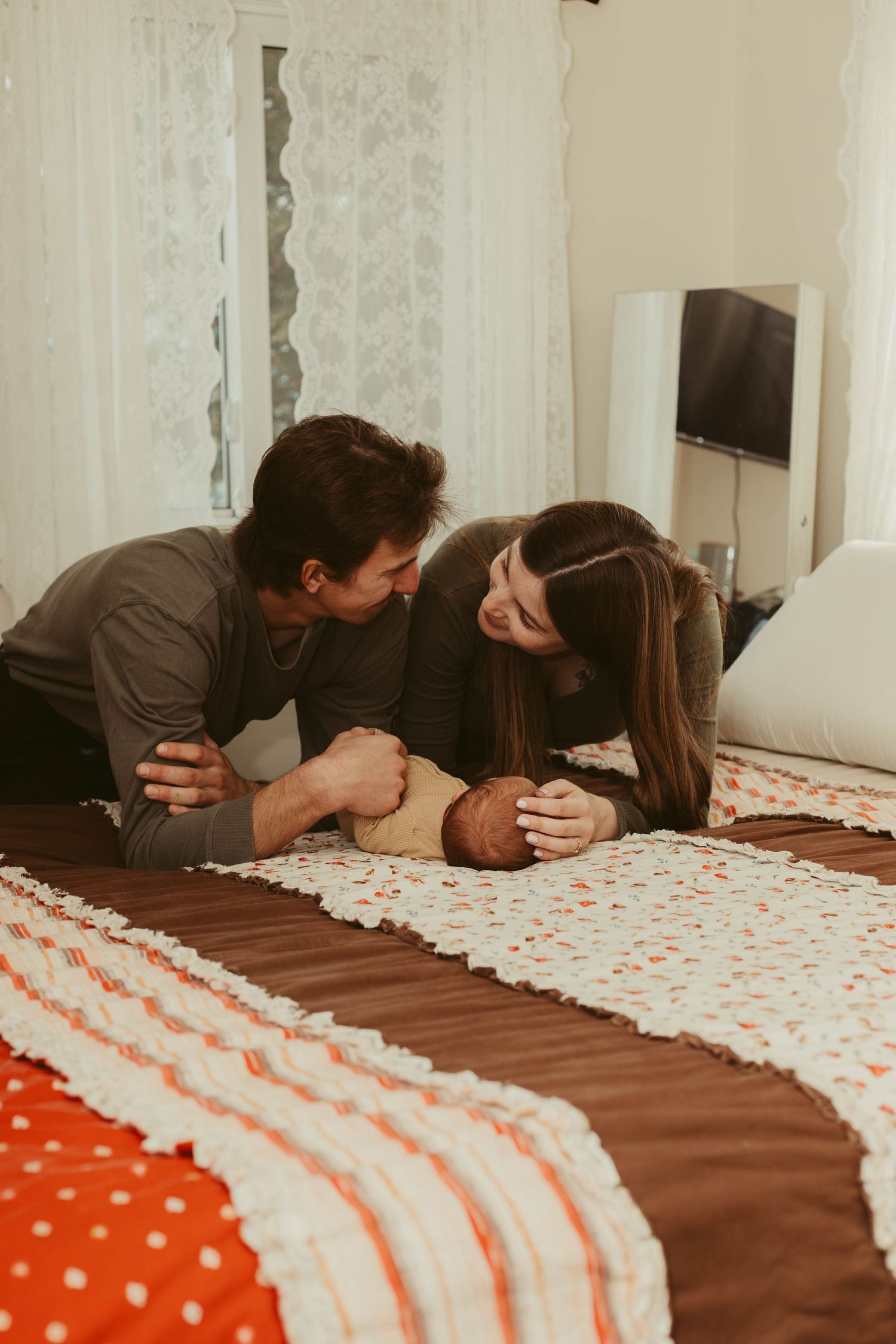 New parents lying on bed gazing at their newborn baby on a colorful patchwork quilt.