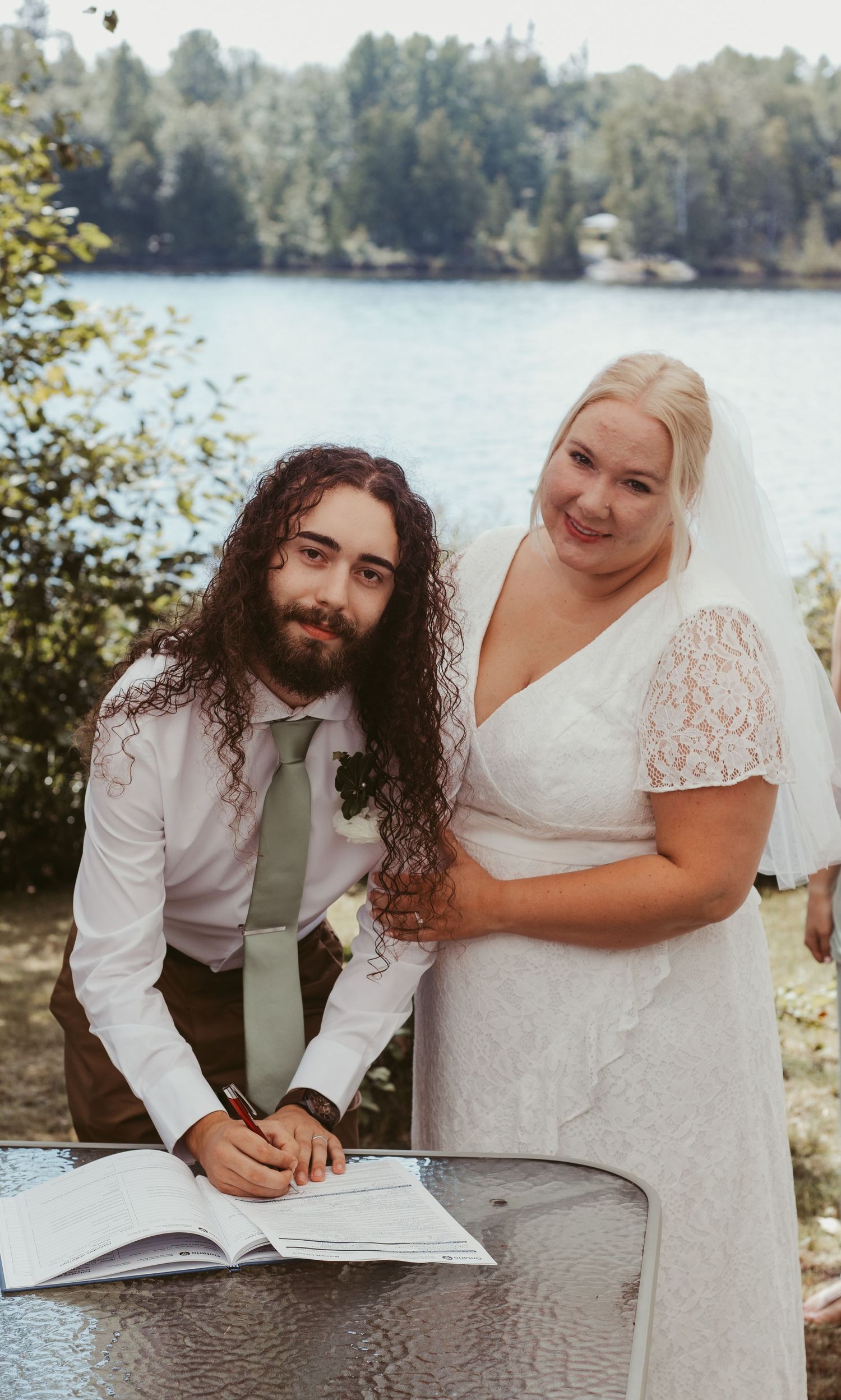 Groom with long curly hair signs wedding registry book beside smiling blonde bride near lakeside setting.