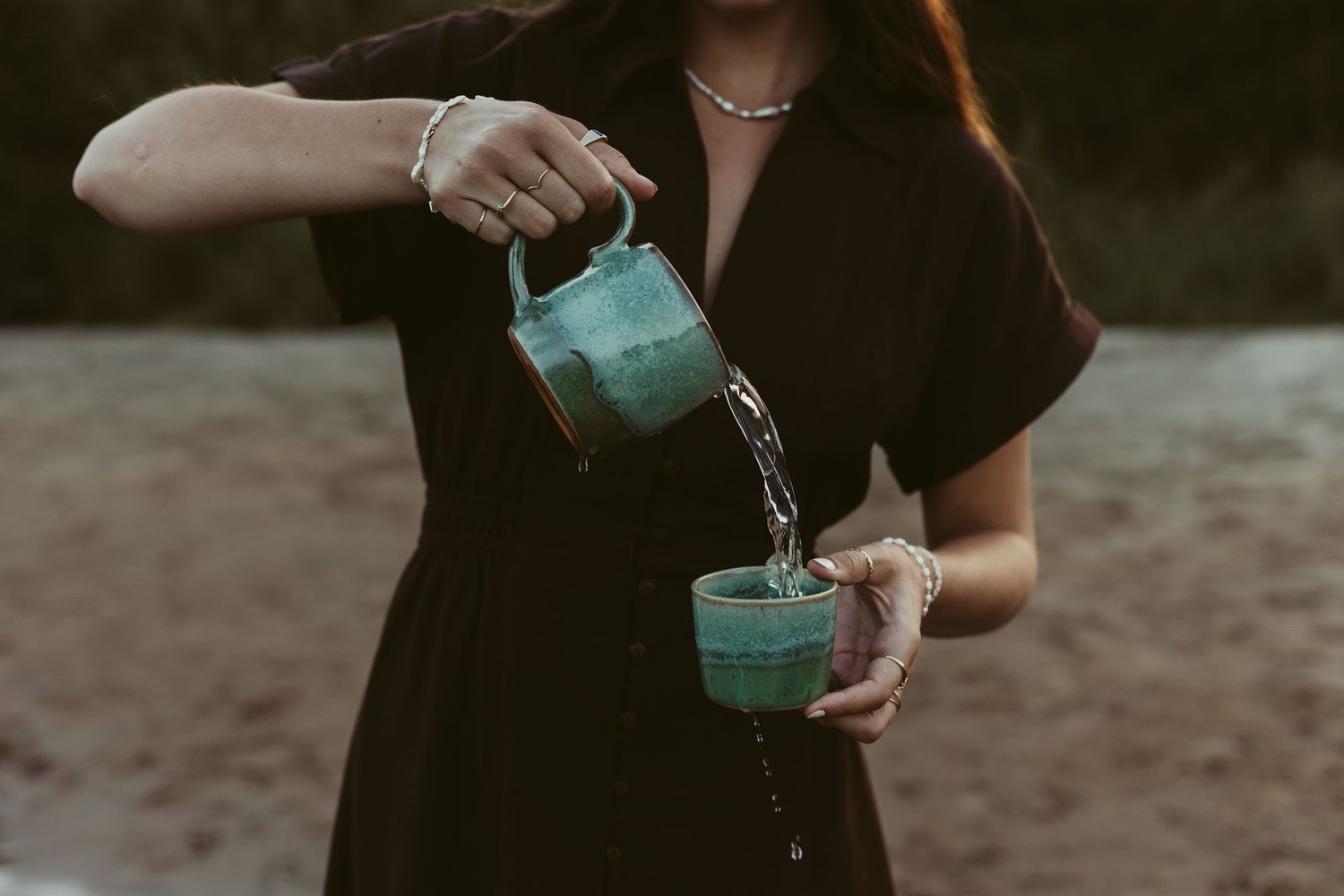 Woman in black dress pouring water from teal ceramic pitcher into matching cup outdoors.