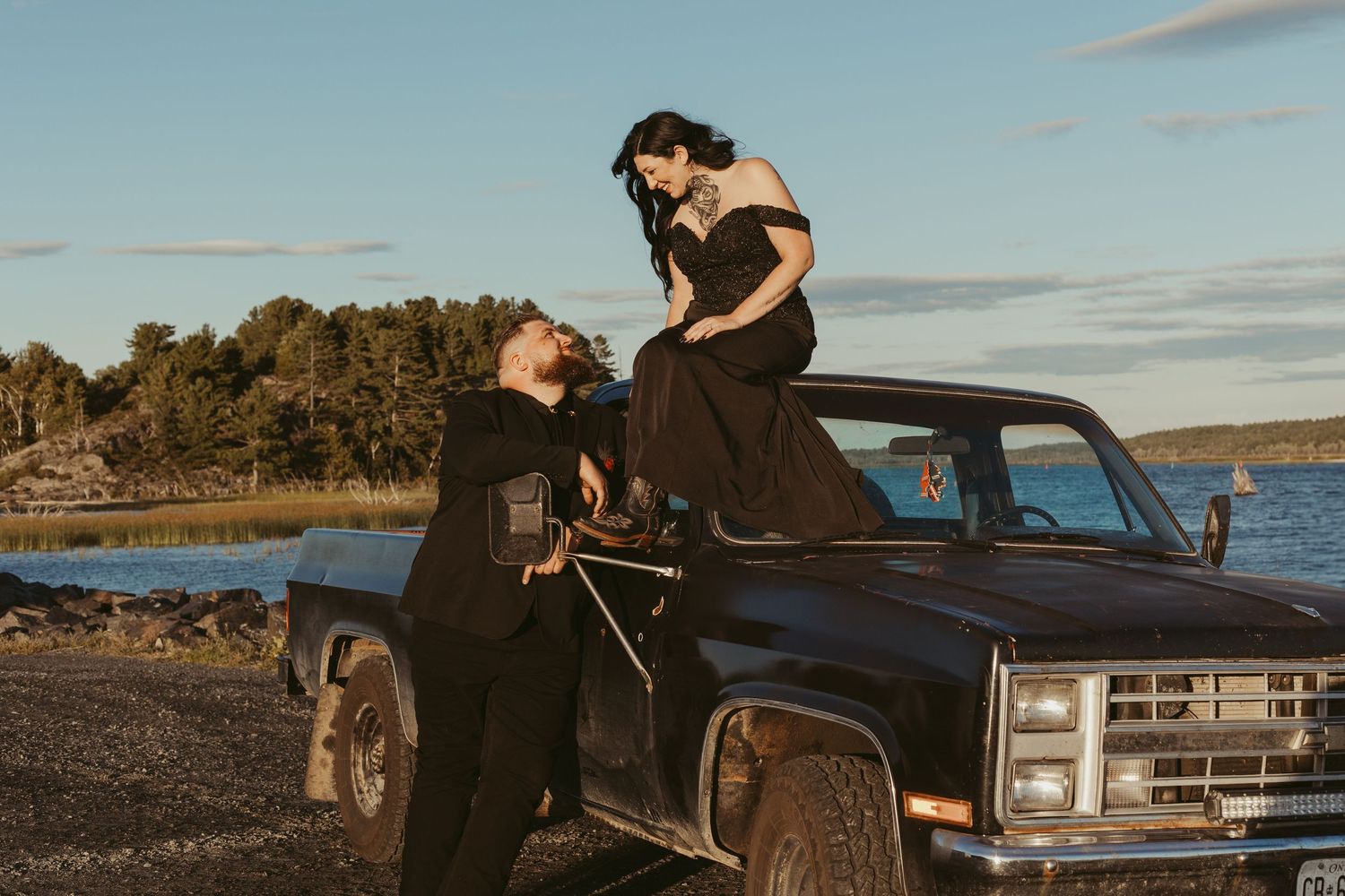 Couple posing with a vintage black truck near a waterfront at golden hour.