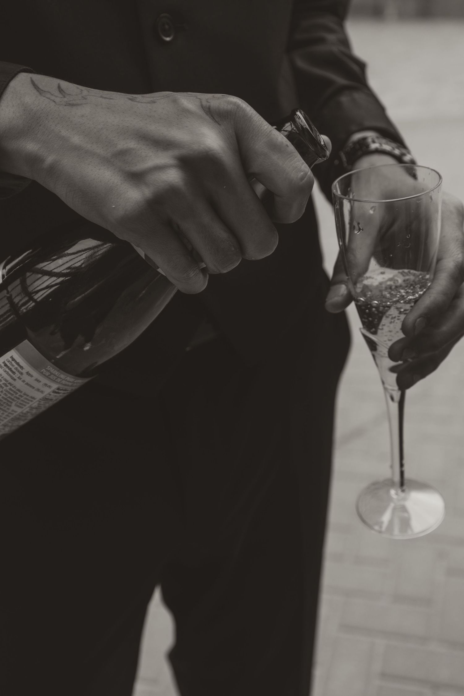 Groom in black suit pouring champagne into crystal flute glass, black and white wedding detail.