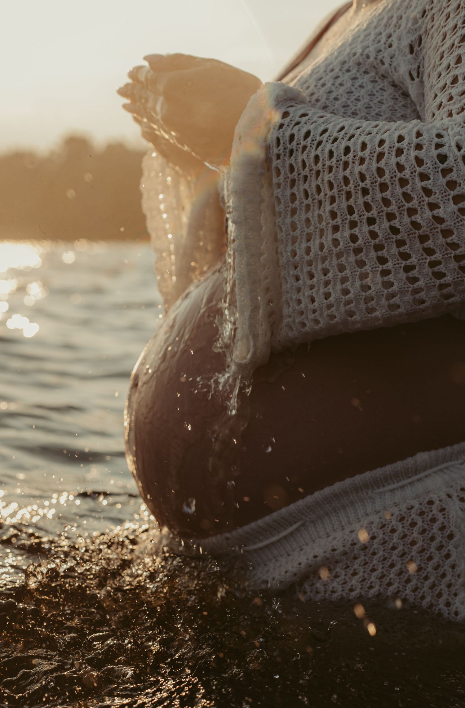 Pregnant woman in white knit top pouring water over baby bump at golden hour by lake.