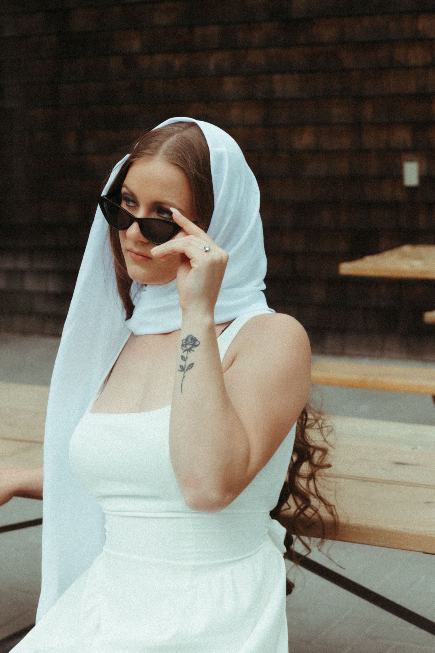 Bride in white dress and veil lowering sunglasses with a rose tattoo on her arm.