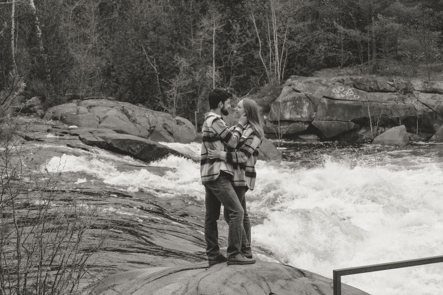 Couple embraces romantically on rocky riverbank beside rushing white water rapids in black and white.