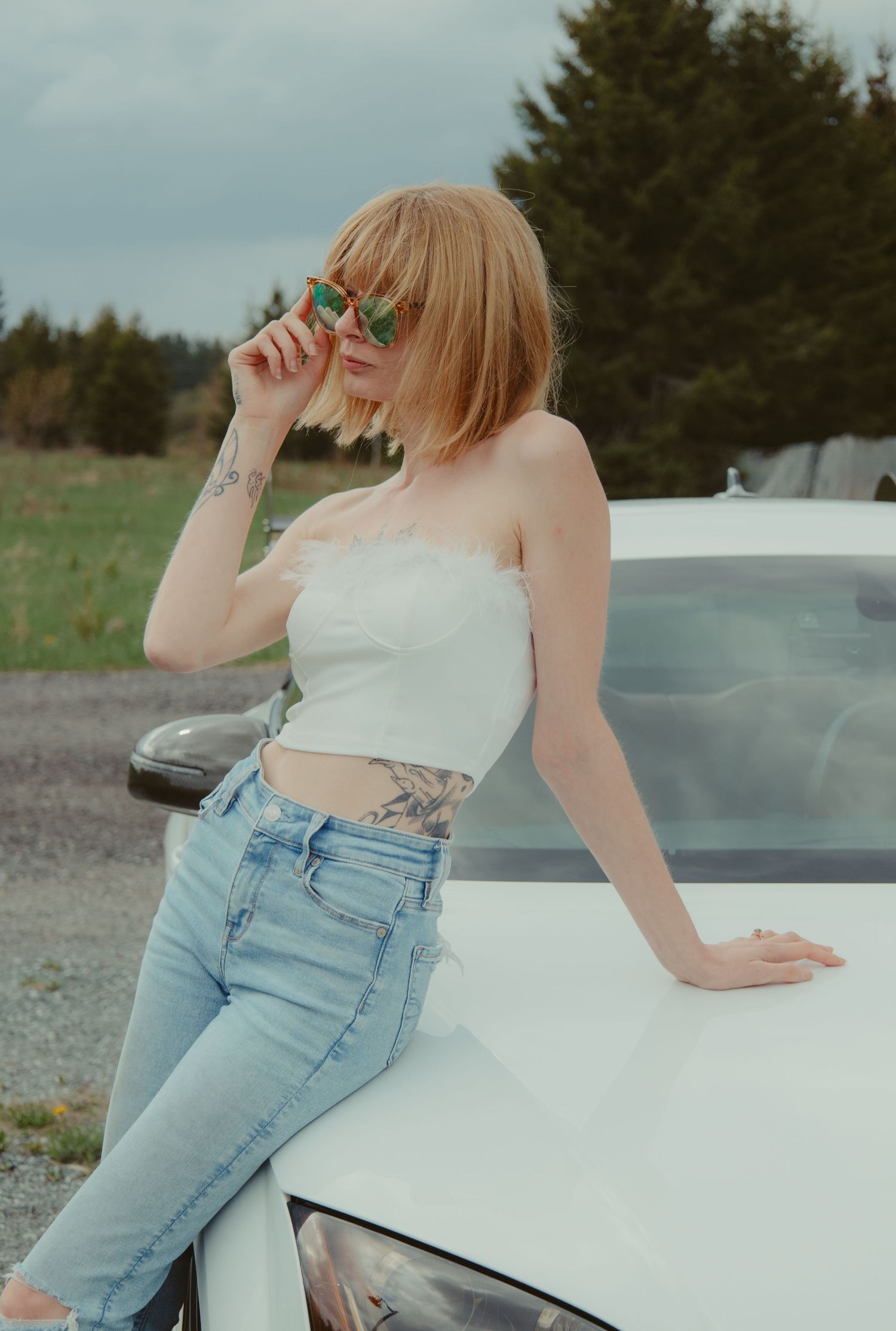 Redhead woman with tattoos in white tube top and jeans leans on white car outdoors.