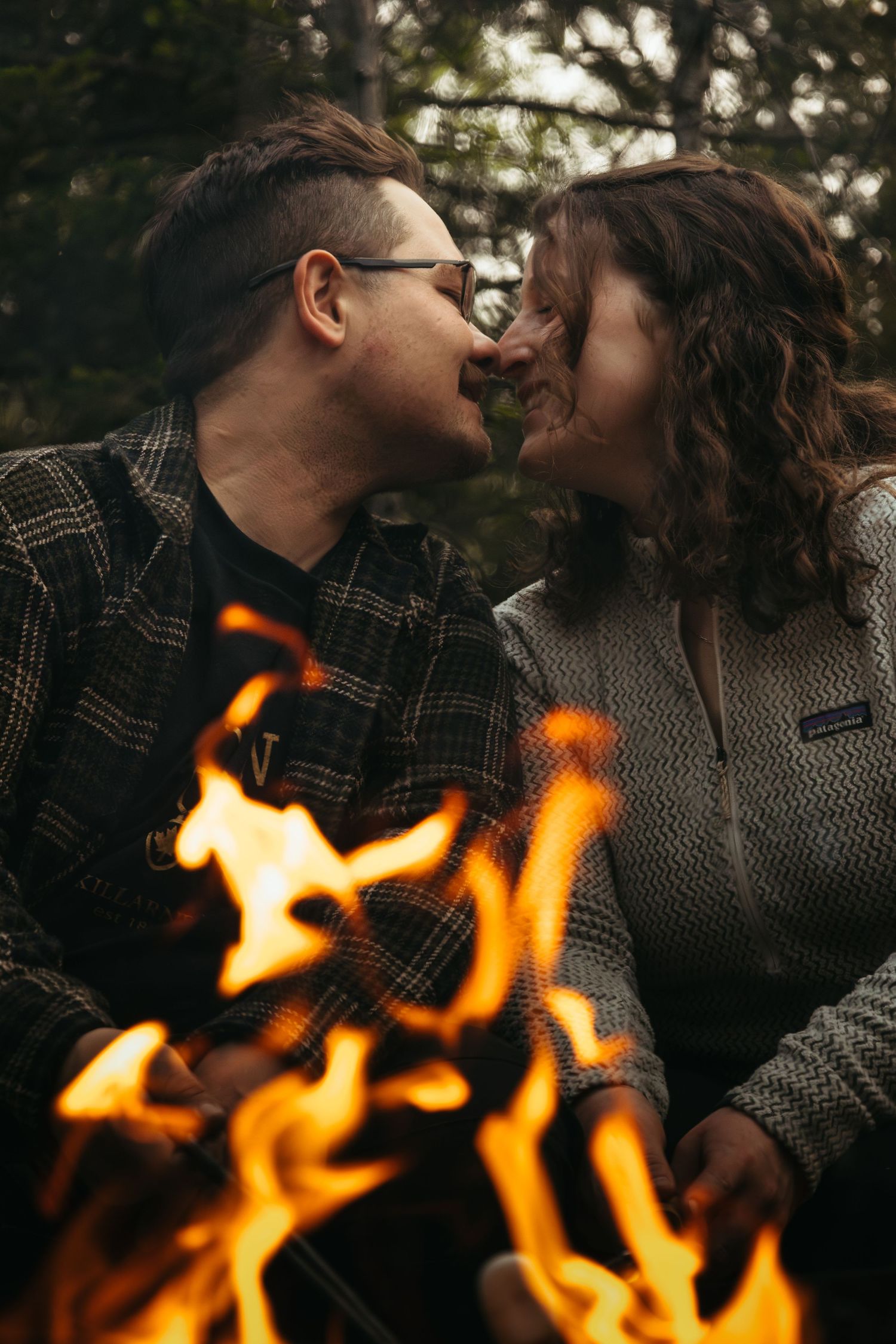 Romantic couple kissing near a campfire with flames in foreground in forest setting.