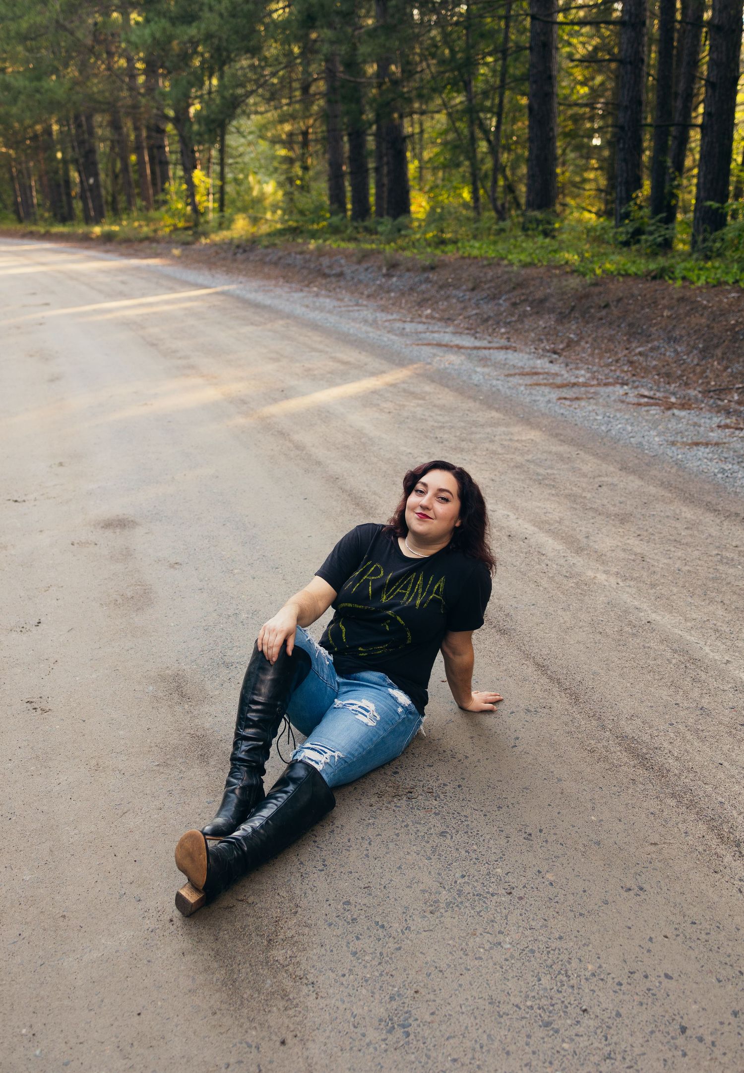 Woman in Nirvana tee, ripped jeans, and tall black boots sitting on a rural road surrounded by lush green forest.