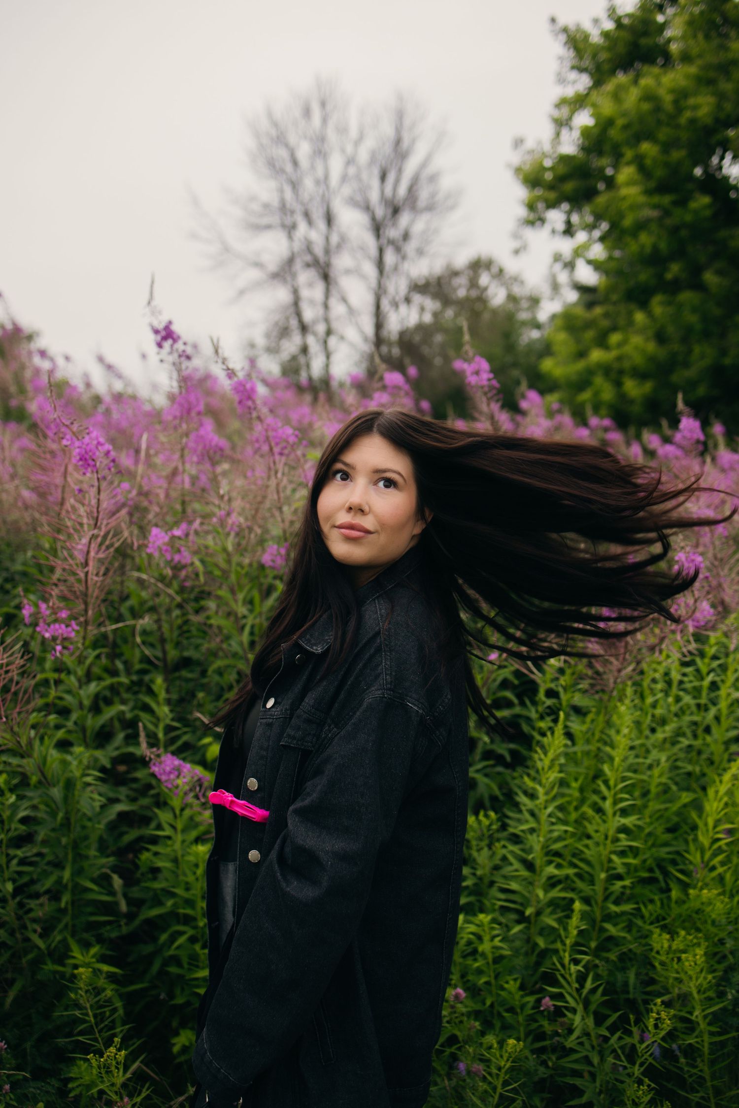 Young woman with flowing dark hair stands among purple wildflowers in a lush green field wearing a black jacket.