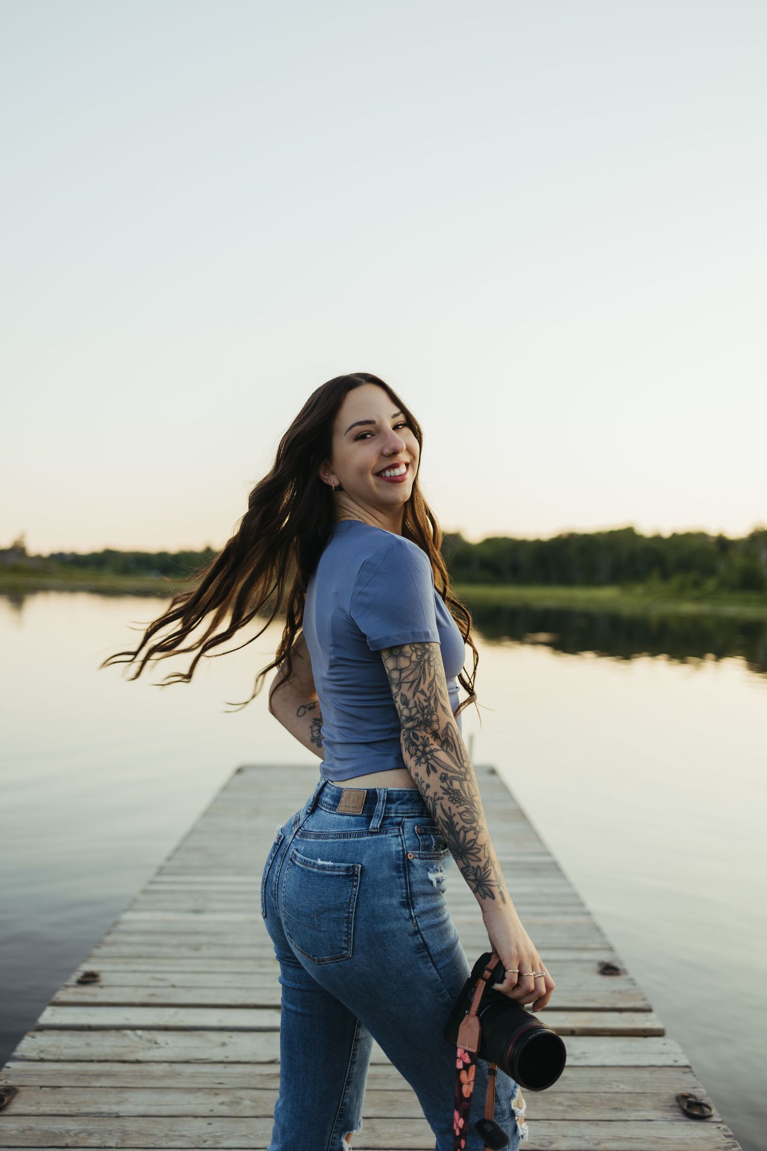 Smiling tattooed woman in blue crop top and jeans holding camera on wooden dock by lake.