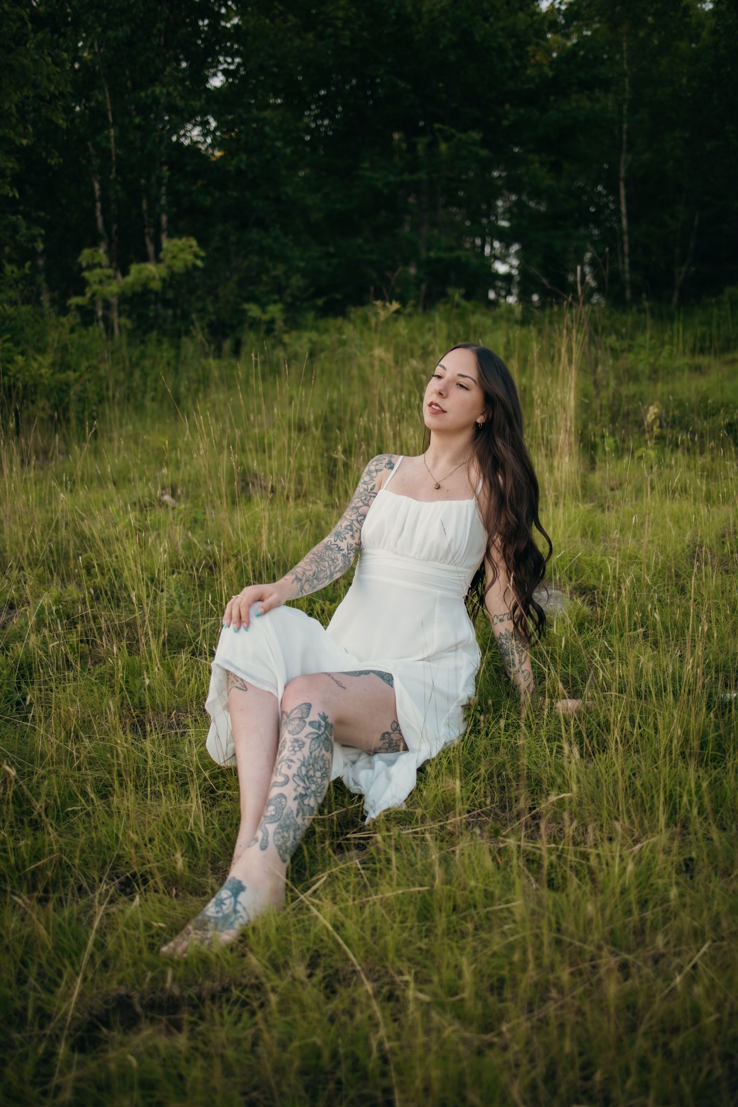 Woman with tattoos in white dress sitting in green meadow with forest background.