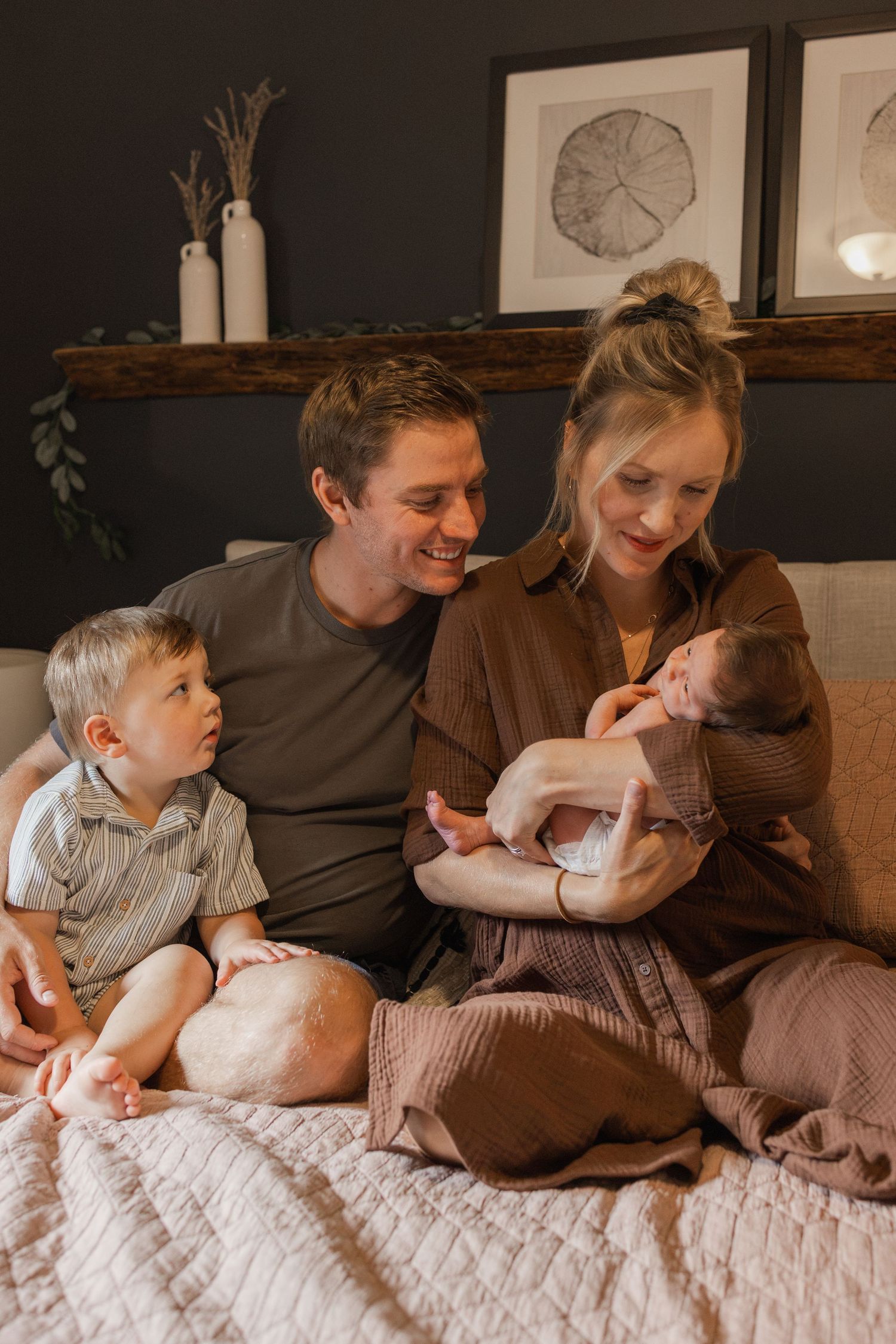 A happy family of four sits on a bed, with parents smiling at their newborn baby while a toddler sits beside them.