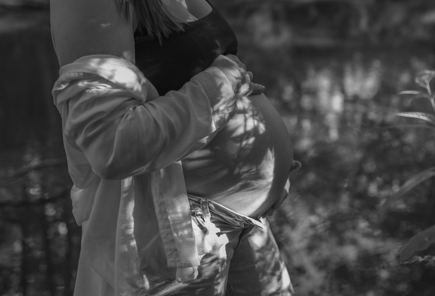 Black and white photo of a pregnant woman cradling her bare baby bump outdoors in dappled sunlight.