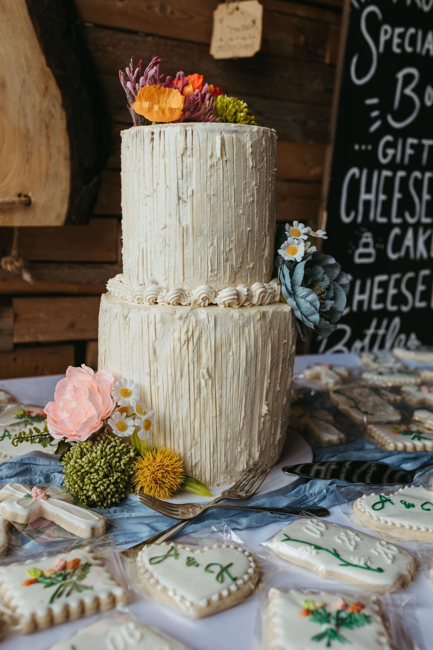 Two-tier rustic textured white wedding cake decorated with colorful flowers, surrounded by decorated sugar cookies on a table.