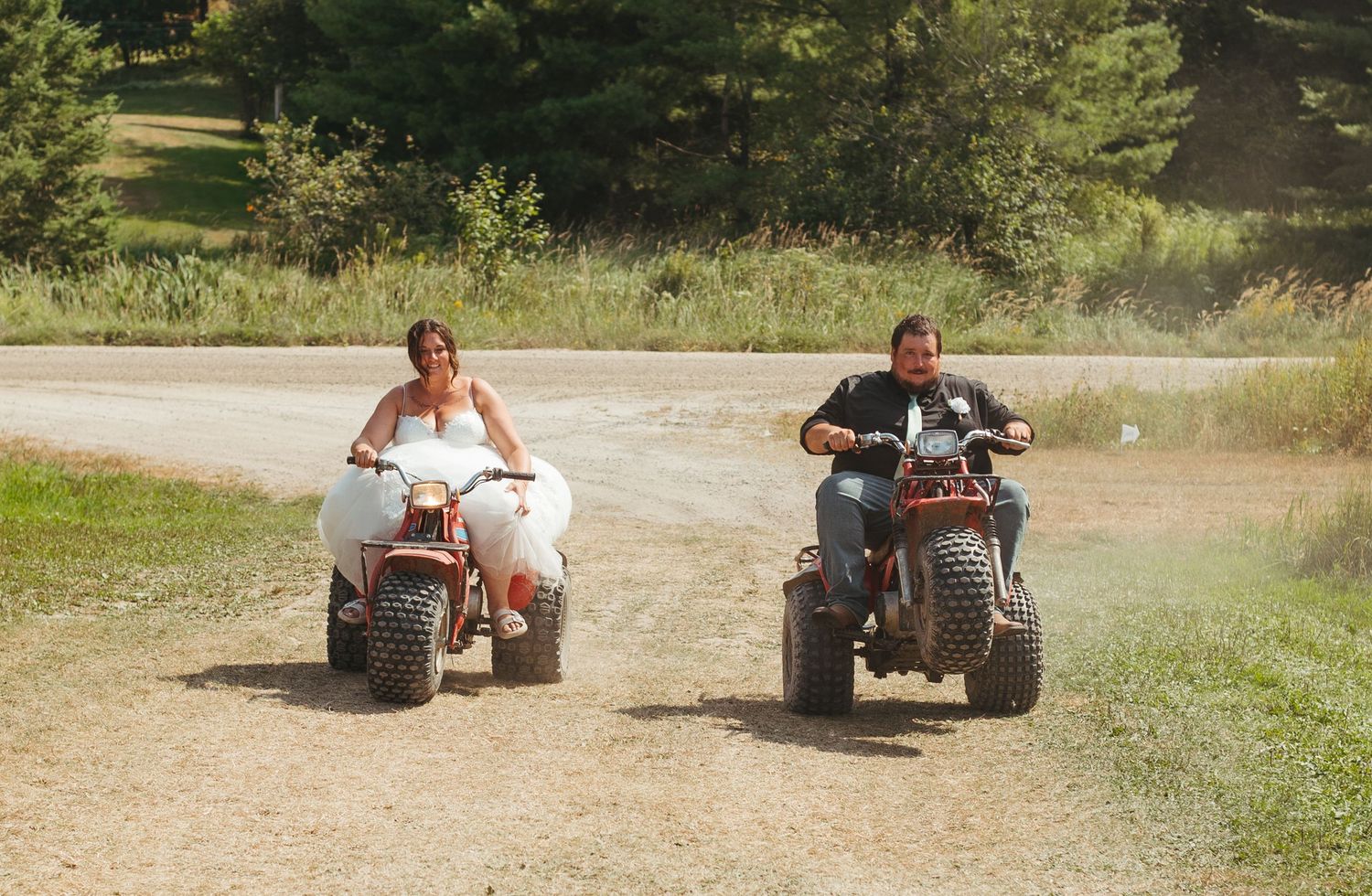 Bride and groom riding ATVs together on dirt path at outdoor wedding.