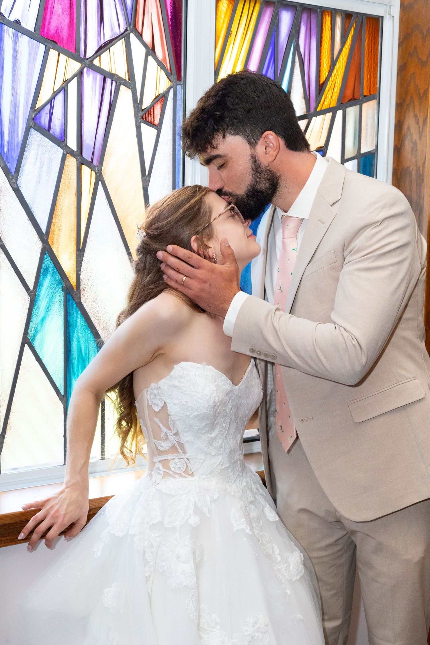 Groom tenderly kisses bride on the forehead before colorful stained glass windows in a church ceremony.