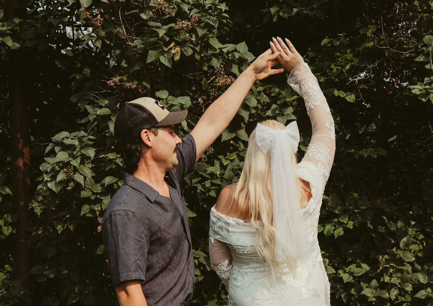 Groom spinning bride in a playful dance outdoors surrounded by lush green trees.