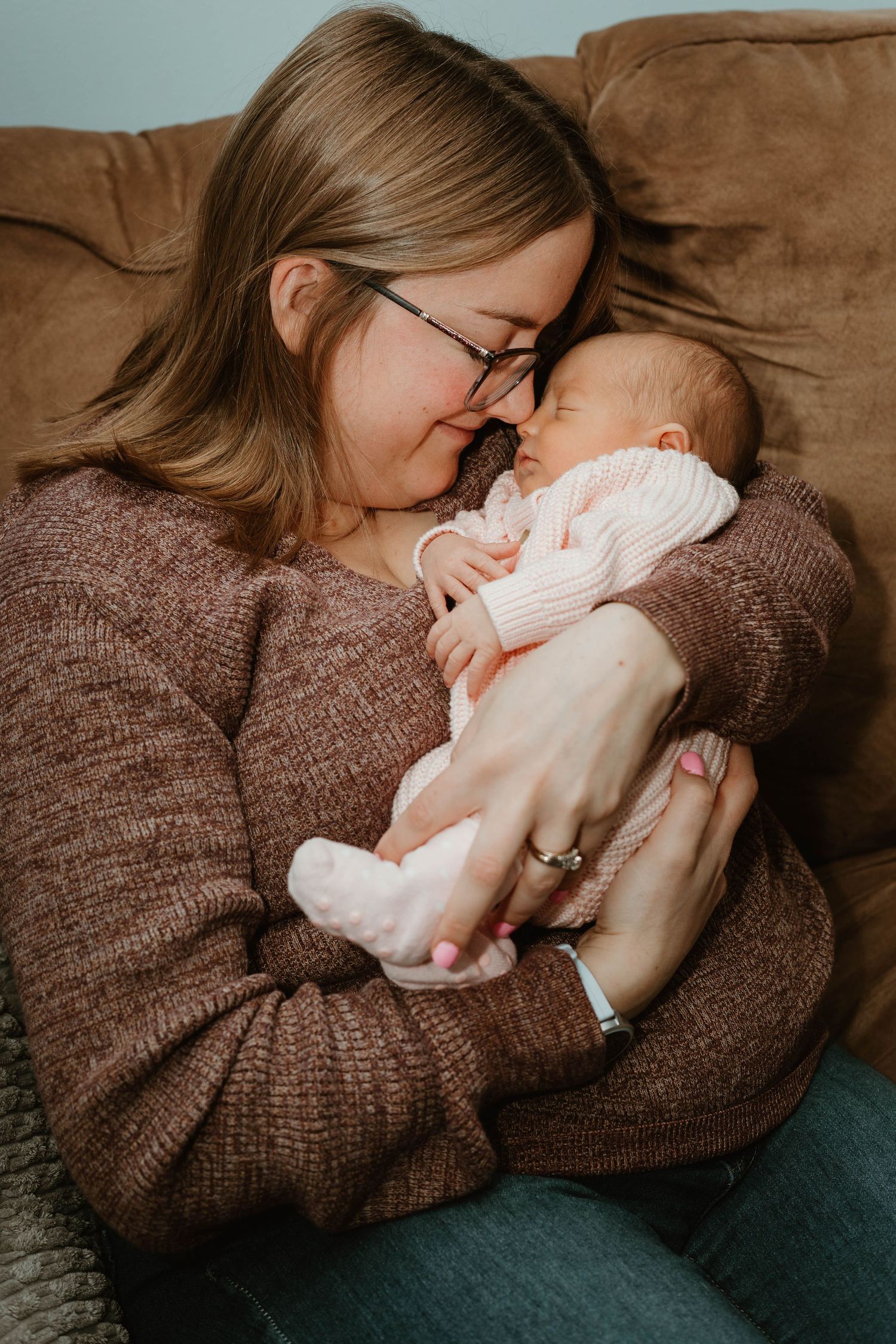 A mother in a brown sweater tenderly holds and nuzzles her sleeping newborn baby dressed in pink.