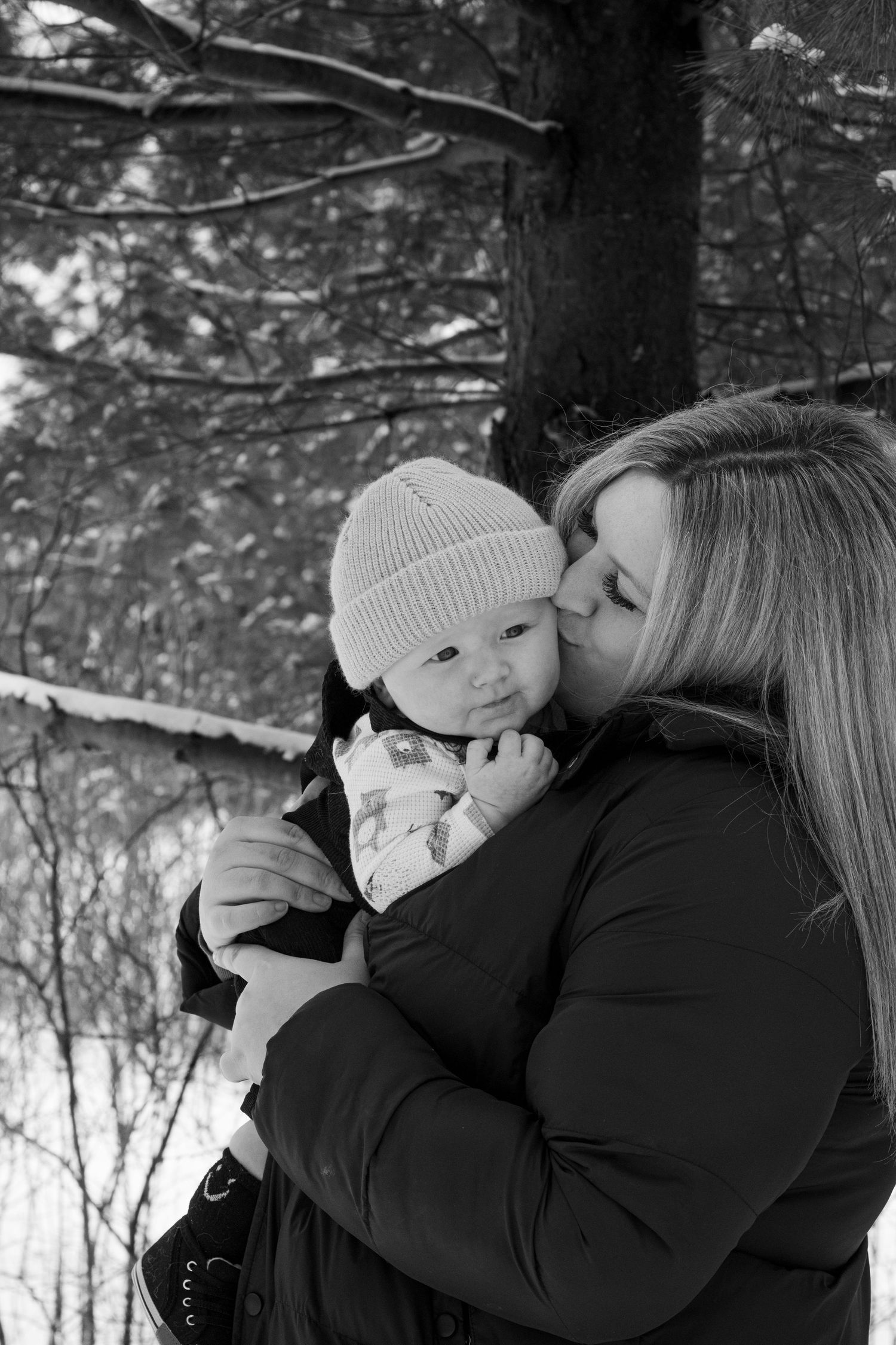 Mother kissing baby in knit hat outdoors in snowy winter forest, black and white photo.